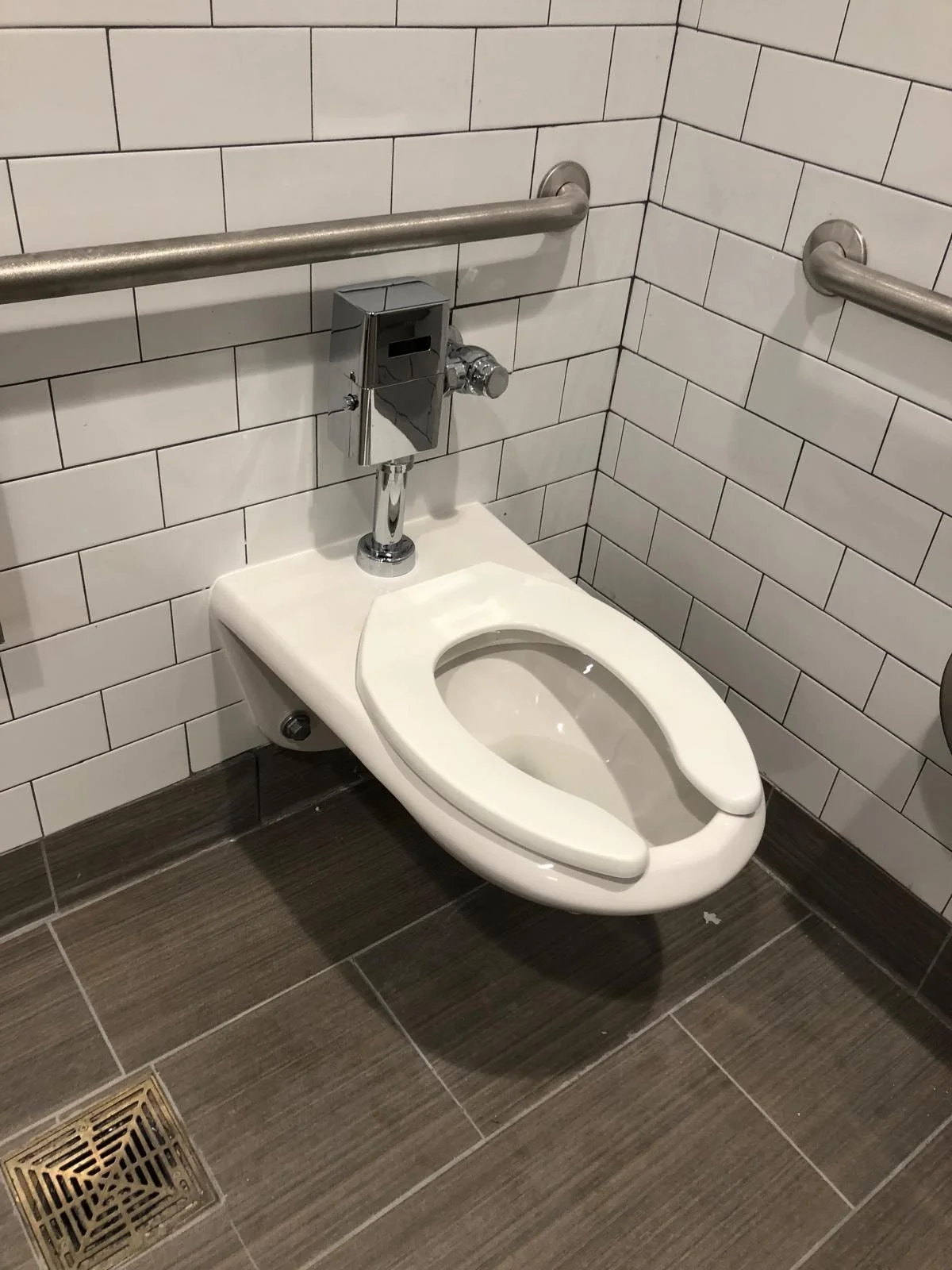A wheelchair-accessible toilet with metal grab bars on the sides in a bathroom with tiled walls and floor.