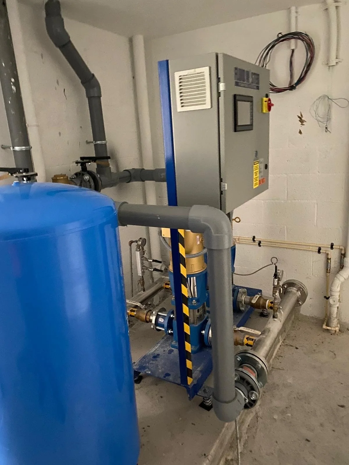 An industrial water system with pipes, a large blue tank, and an electrical control panel mounted on a cinder block wall.