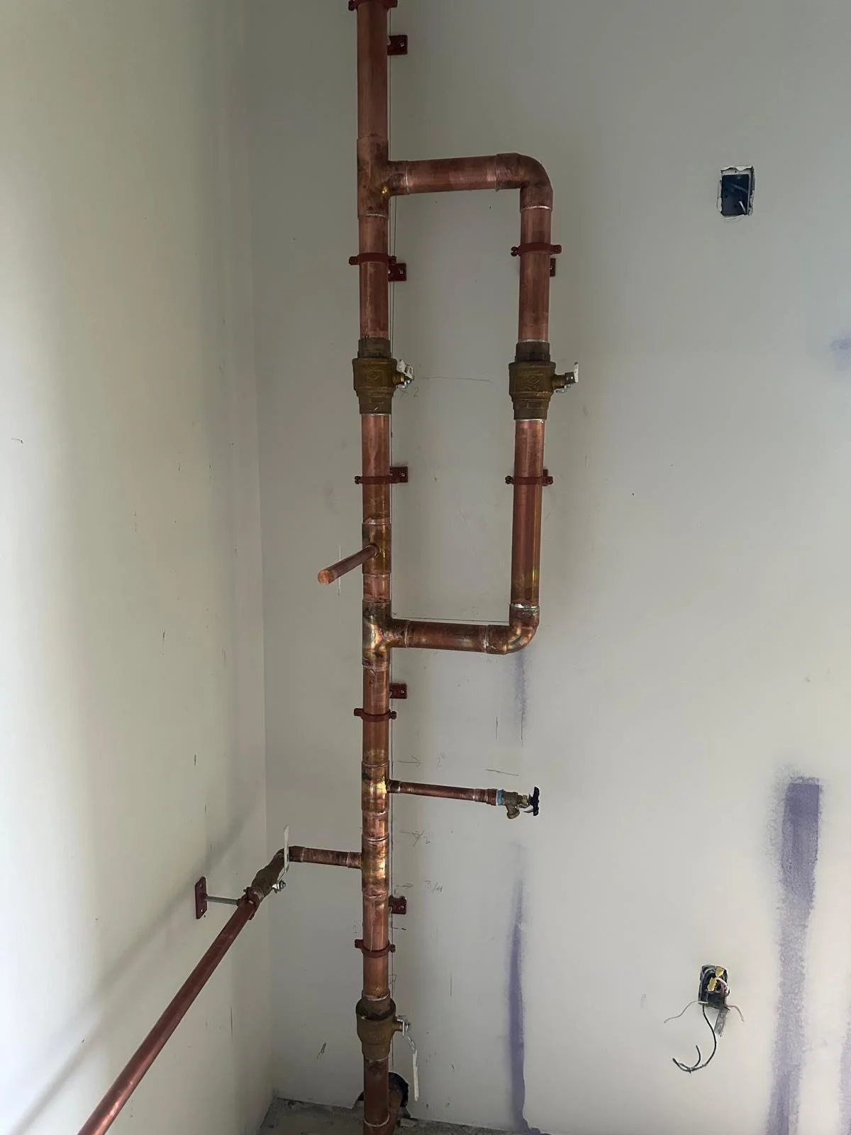 Copper plumbing pipes and valves mounted on a wall, with some wall outlets and wires visible.