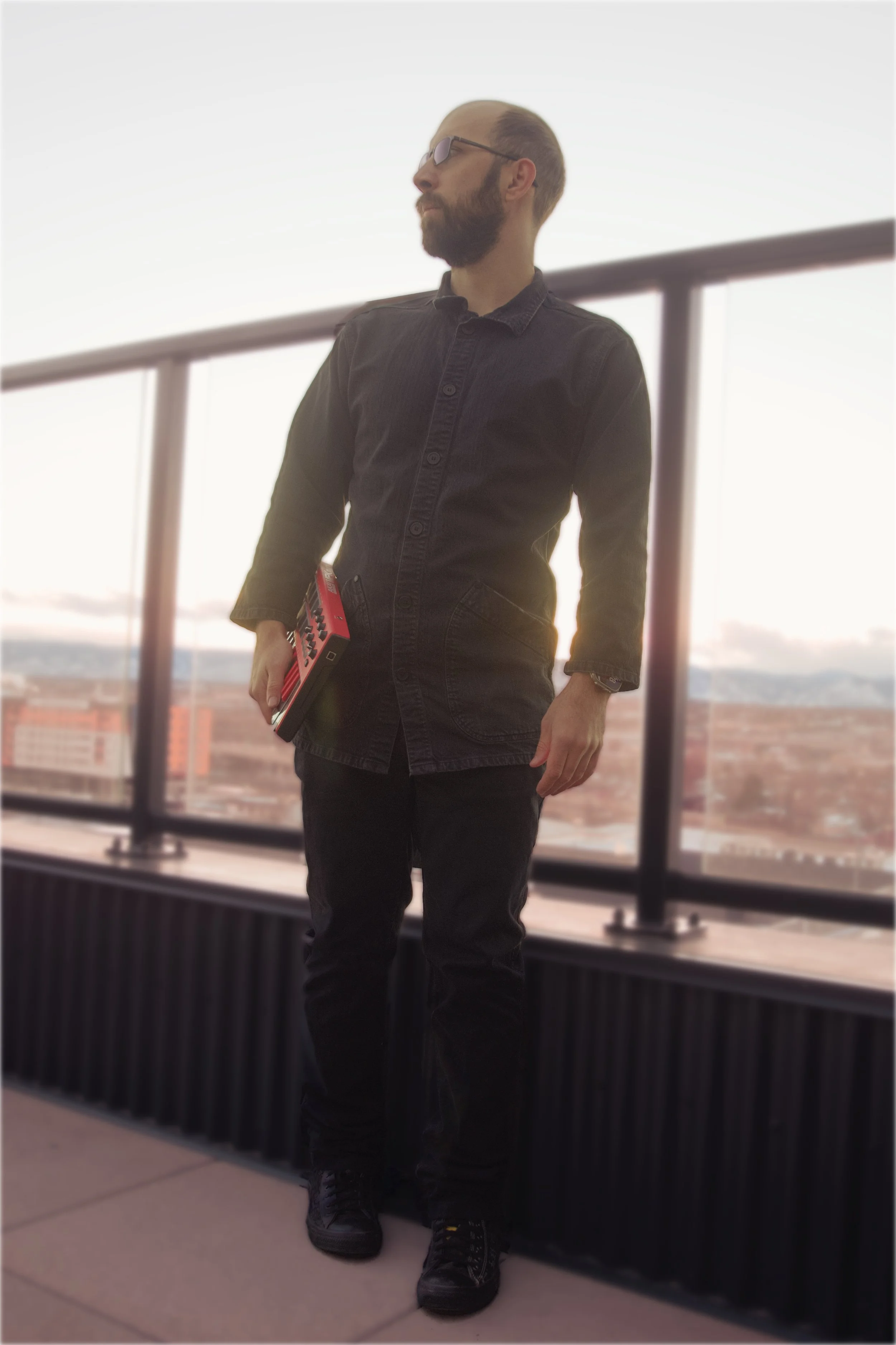 A man with glasses, a beard, and light-colored hair holds a red keyboard and stands on a balcony at sunset, overlooking a cityscape.