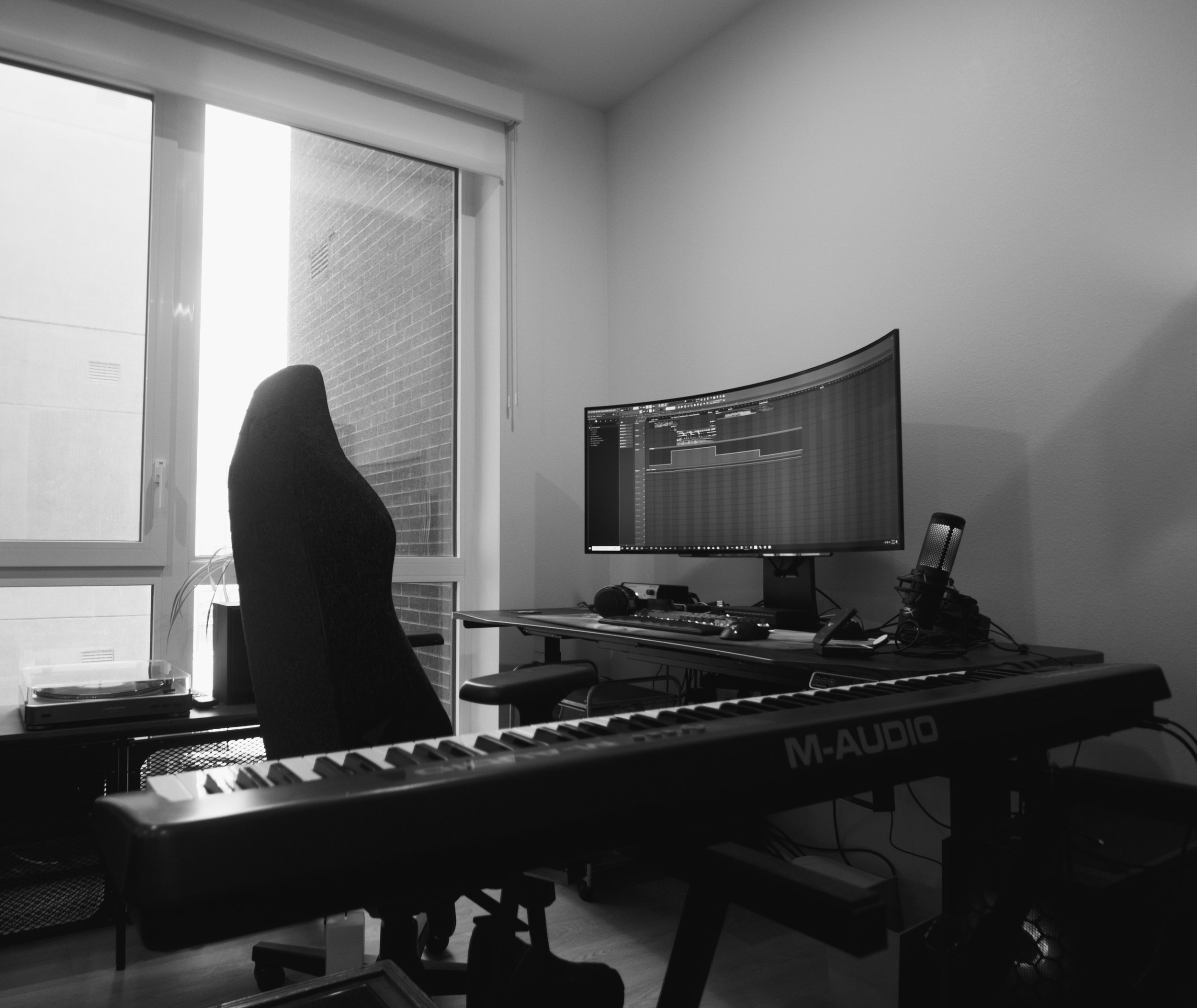 A home studio setup with a curved monitor on a desk, a microphone on a stand, an M-Audio keyboard in the foreground, an office chair, and a window with a roller blind, with a brick exterior wall visible outside.