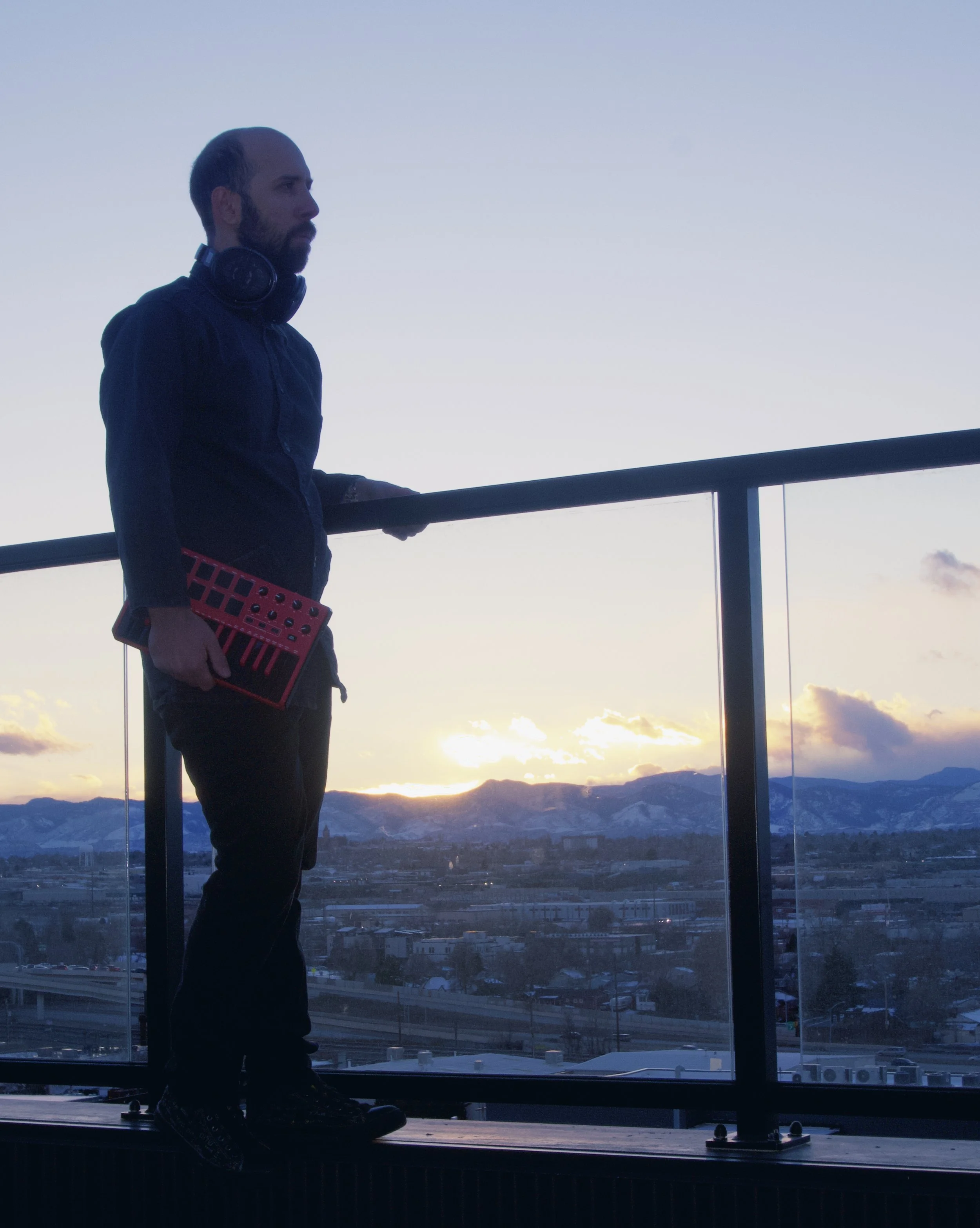 A man with a beard and headphones around his neck standing on a balcony overlooking a city and mountains during sunset, holding a red electronic music controller.