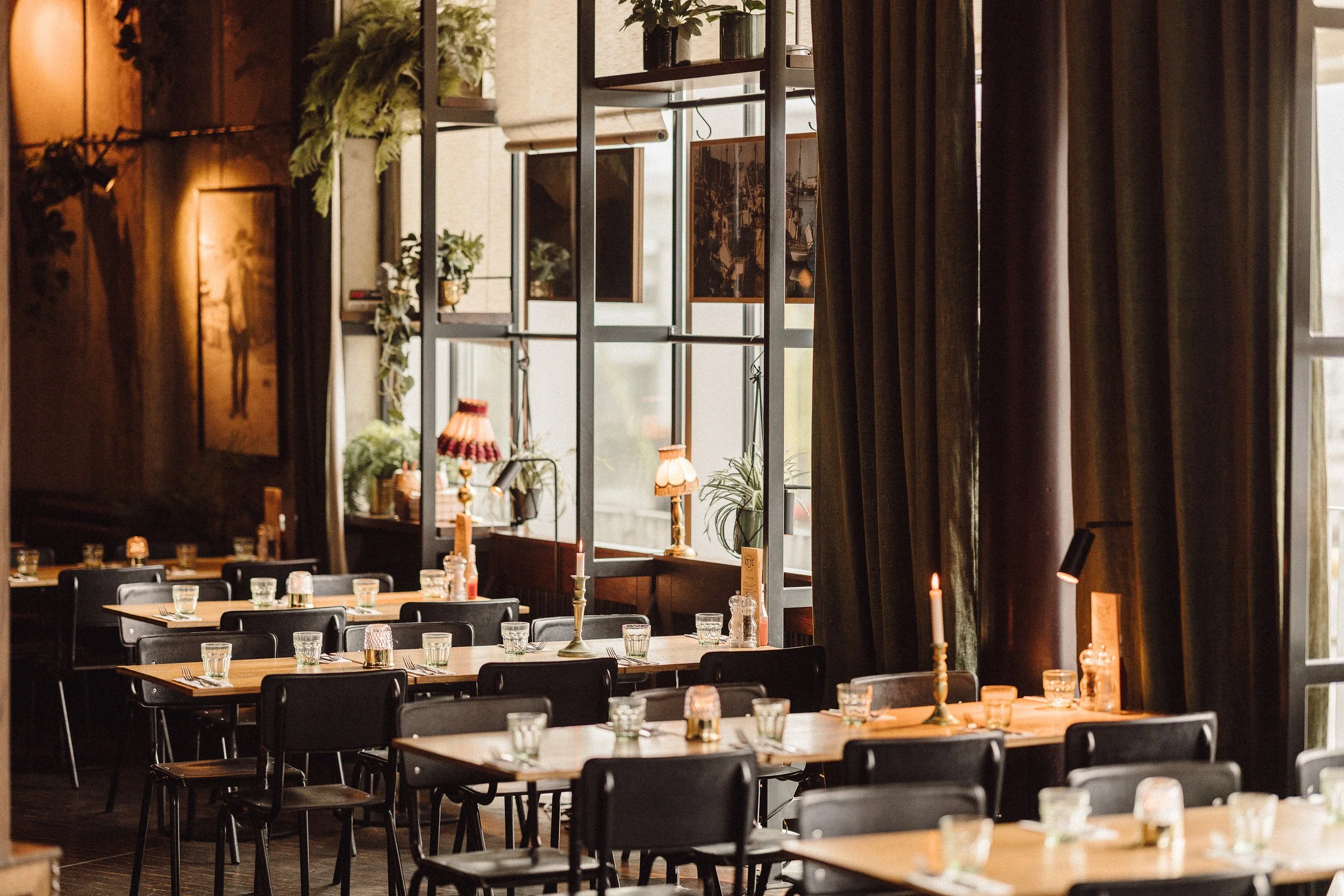 A cozy restaurant interior with wooden tables and black chairs, decorated with candles, glassware, and small plants; large windows with dark drapes allowing natural light to fill the space.