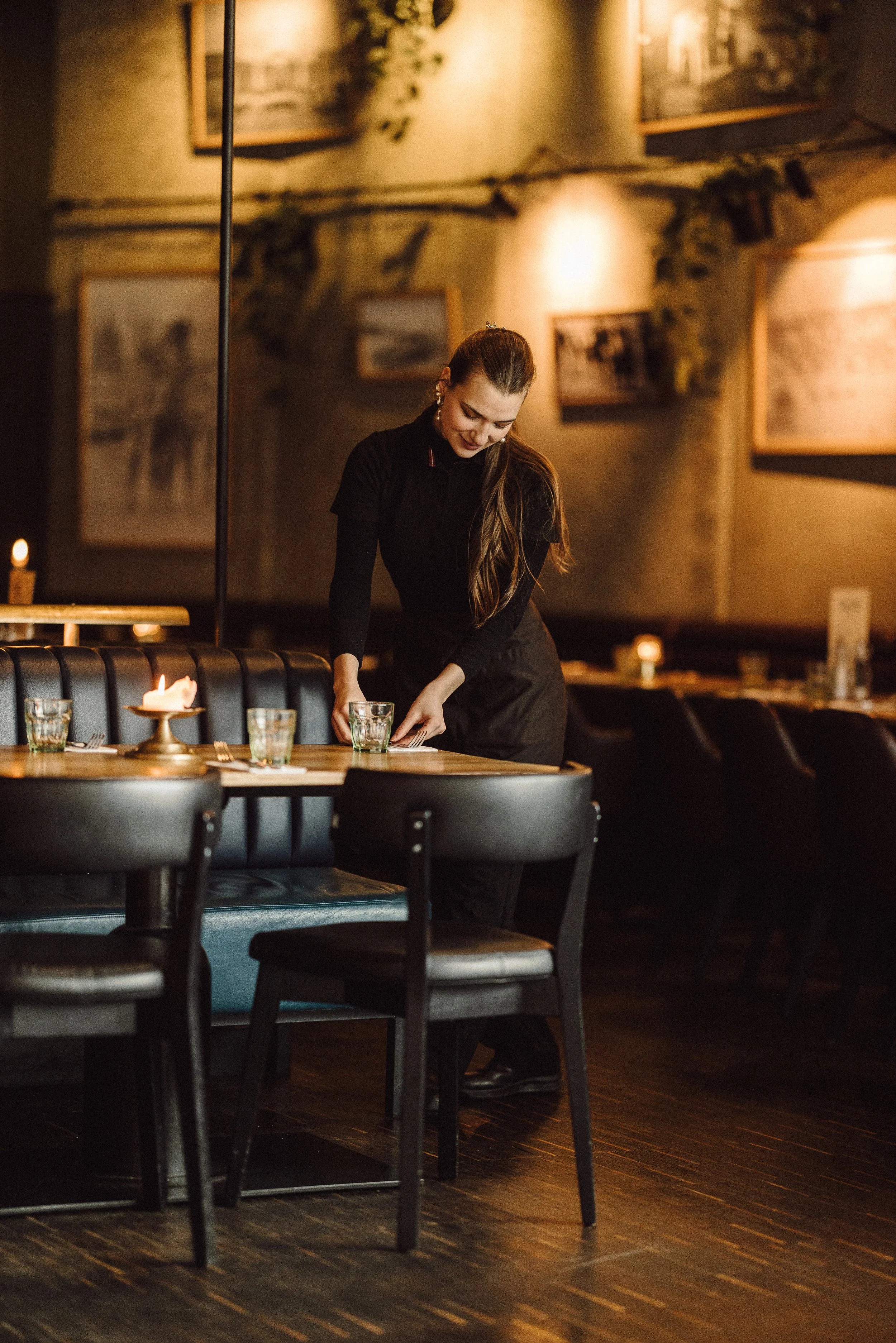 A smiling woman with long hair, dressed in black, is setting a table in a warmly lit restaurant. The table has glasses and candles. There are framed pictures on the wall and candles on other tables in the background.