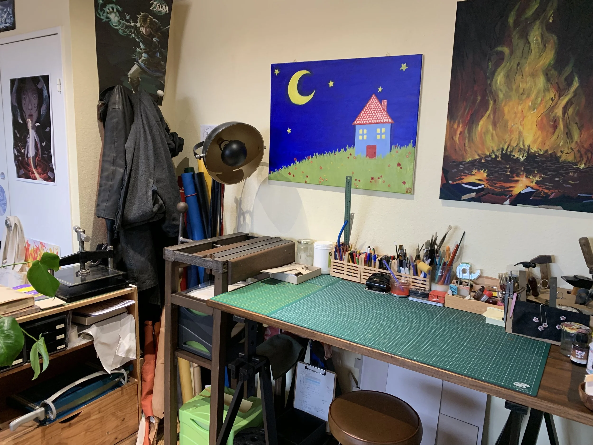 Un atelier d'art avec une grande table de travail recouverte d'un tapis de coupe, divers outils et fournitures d'art, des peintures, et des dessins sur le mur, notamment une maison sous la lune et des montagnes en feu.