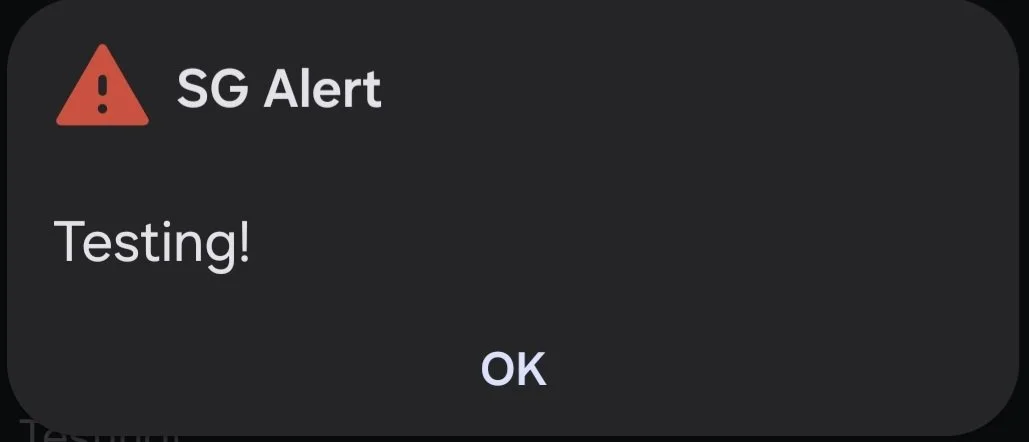 Mobile phone CMAS alert message with a red warning icon, labeled 'SG Alert', stating 'Testing!' with an 'OK' button.