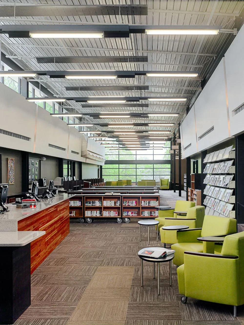 Interior view of a modern library with green chairs, bookshelves, and a high ceiling with exposed metal beams and large windows.