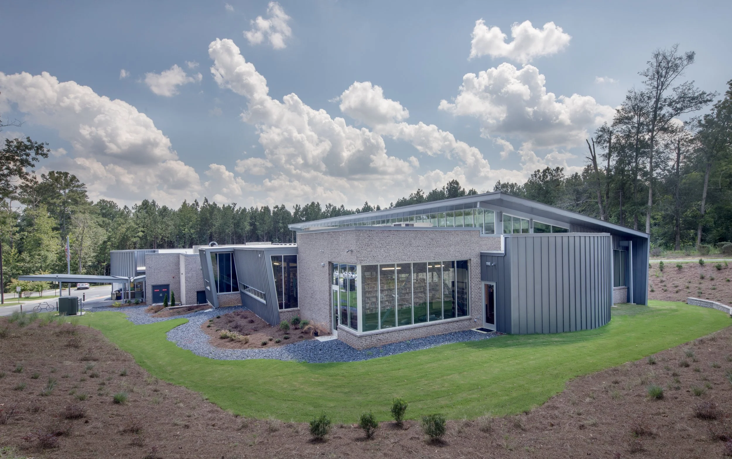 Modern building with large glass windows and metal siding, surrounded by a landscaped area with grass, small trees, and a gravel pathway, under a partly cloudy sky with trees in the background.
