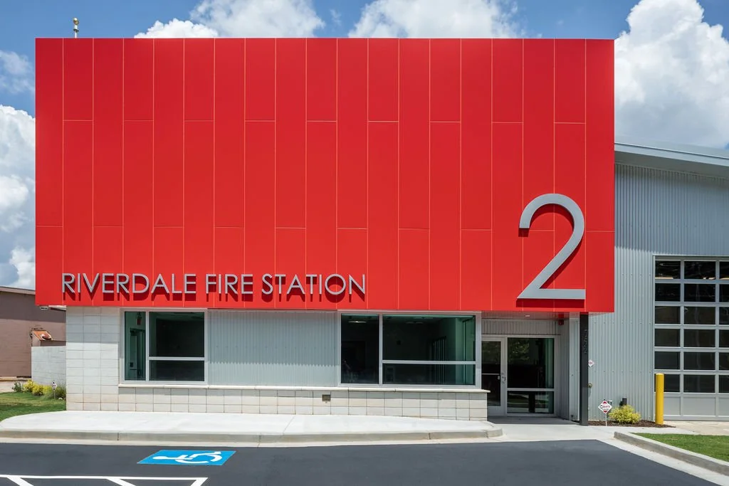 Modern fire station labeled 'Riverdale Fire Station 2' with a large red facade and a glass entrance, located beside a paved parking area with a handicapped spot.