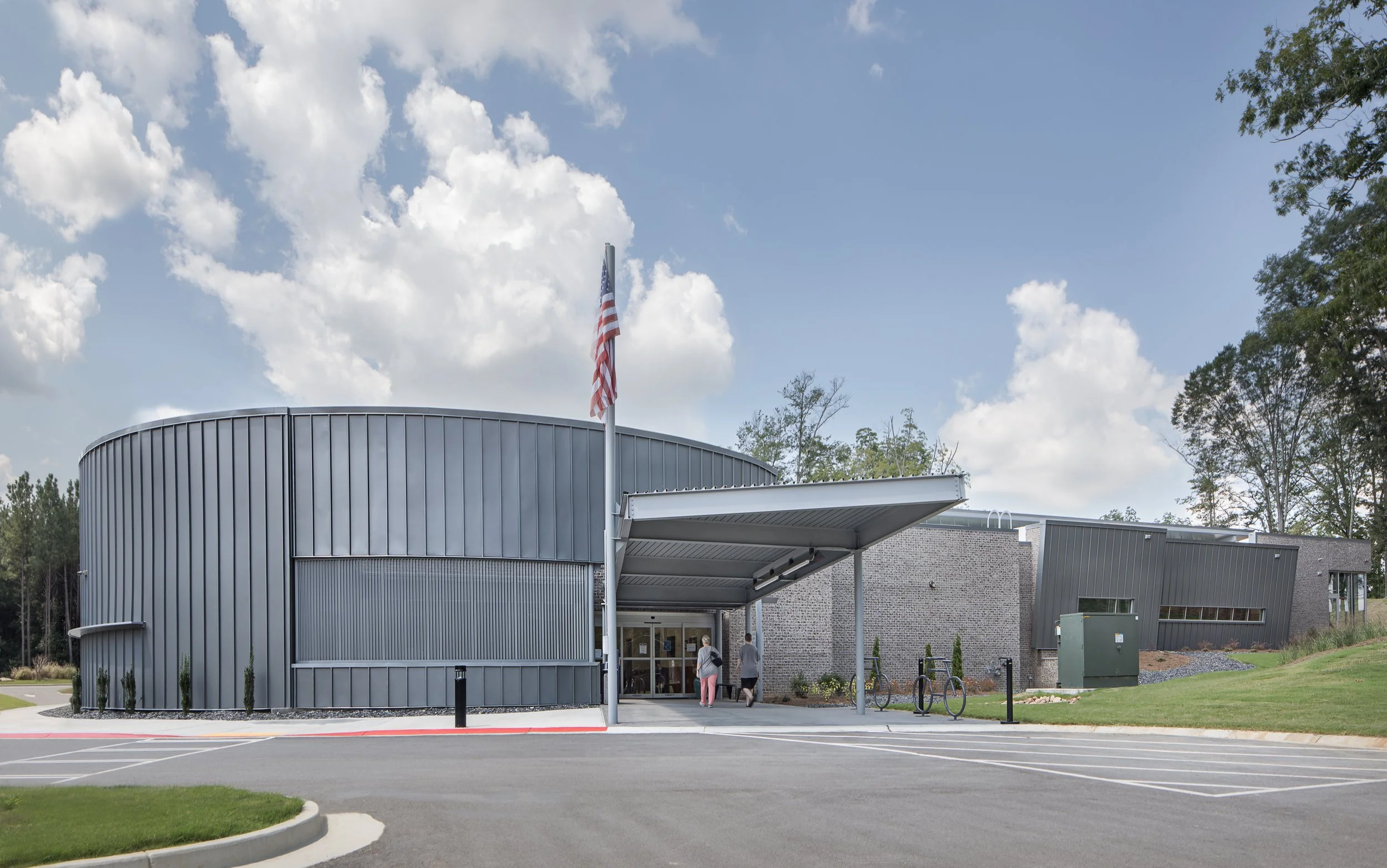 Modern building with metallic siding, an entrance canopy, and an American flag on a flagpole outside. A few people are walking near the entrance, and there is a parking lot with a few parked bikes and a utility box nearby. The sky is partly cloudy wi