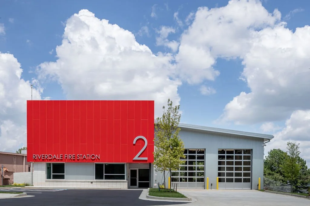 Exterior view of Riverdale Fire Station 2 with a red facade and large glass garage doors, under a partly cloudy sky.