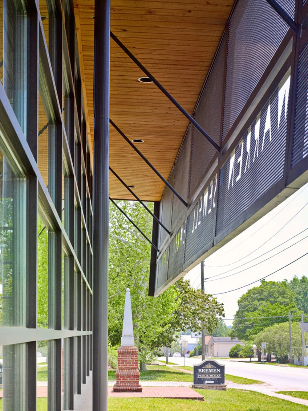 Exterior view of a modern building with large glass windows, metal framework, and a wooden ceiling. A sign reads 'Bremen Public Works,' with trees and a brick monument in the background.