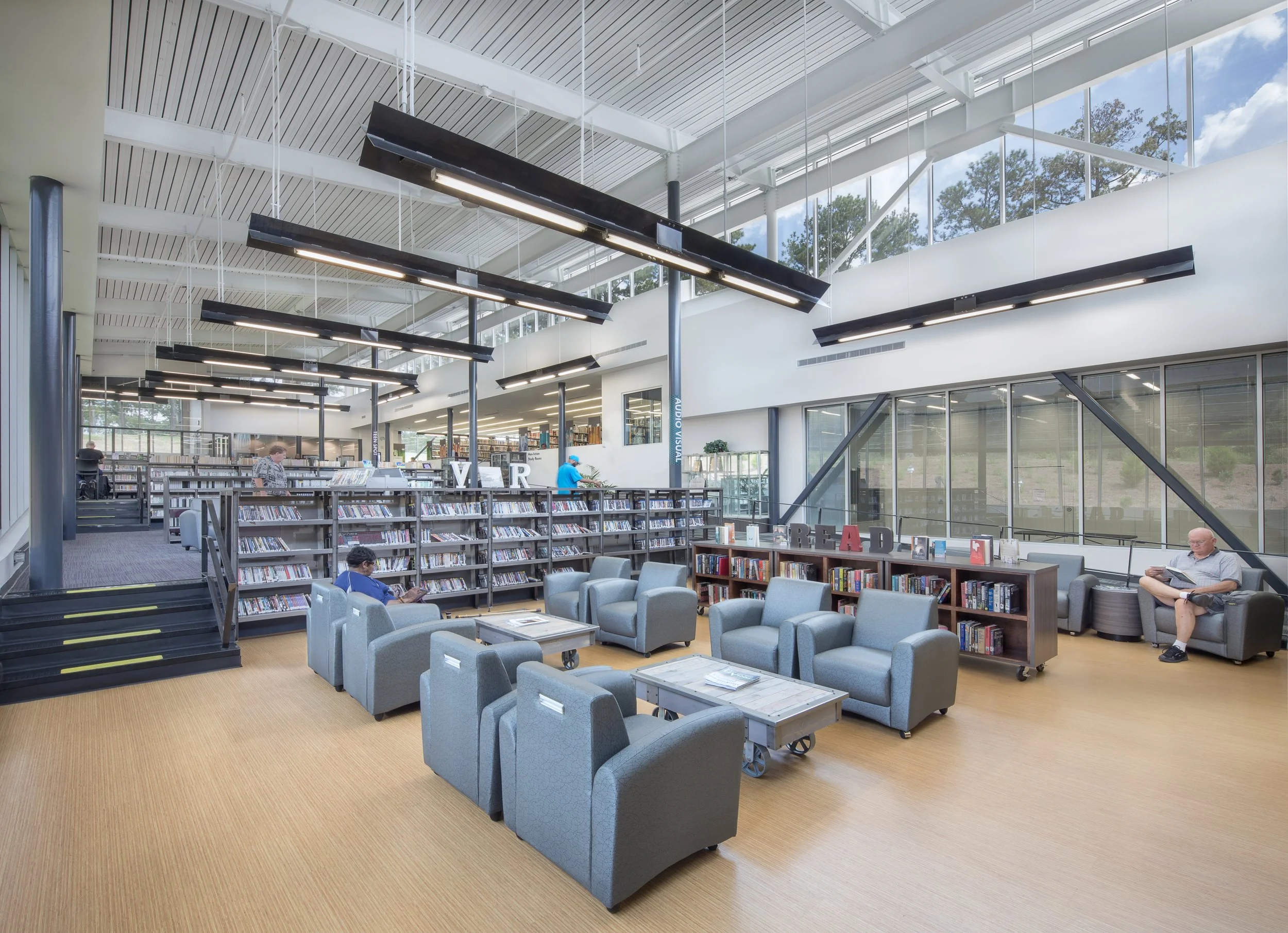 Interior of a modern library with bookshelves, seating areas, and large windows showing trees outside.