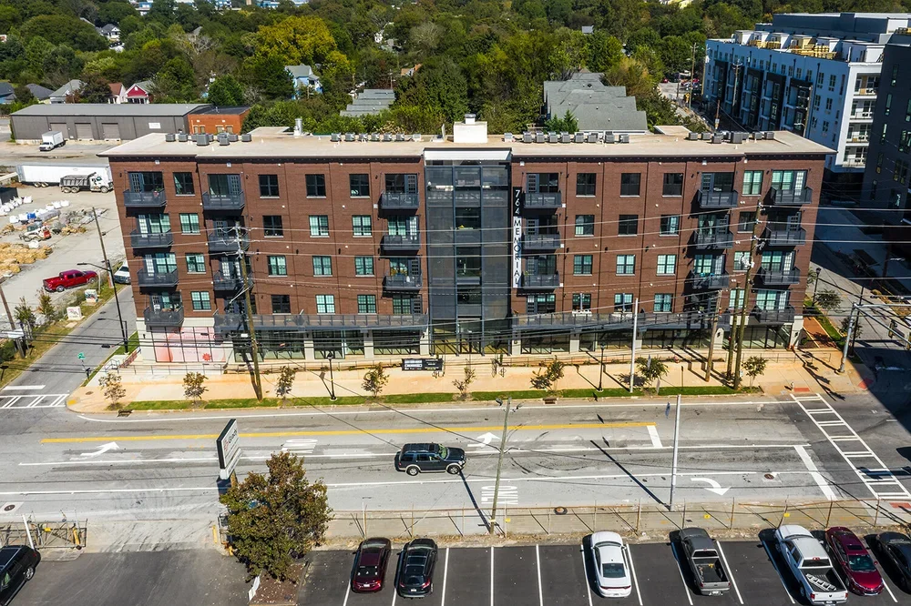 A multi-story residential building with brick exterior, balconies, and a sign that reads '764 Memorial.' The building faces a street with parking spaces and a few cars, with trees planted along the sidewalk. There are residential and commercial struc