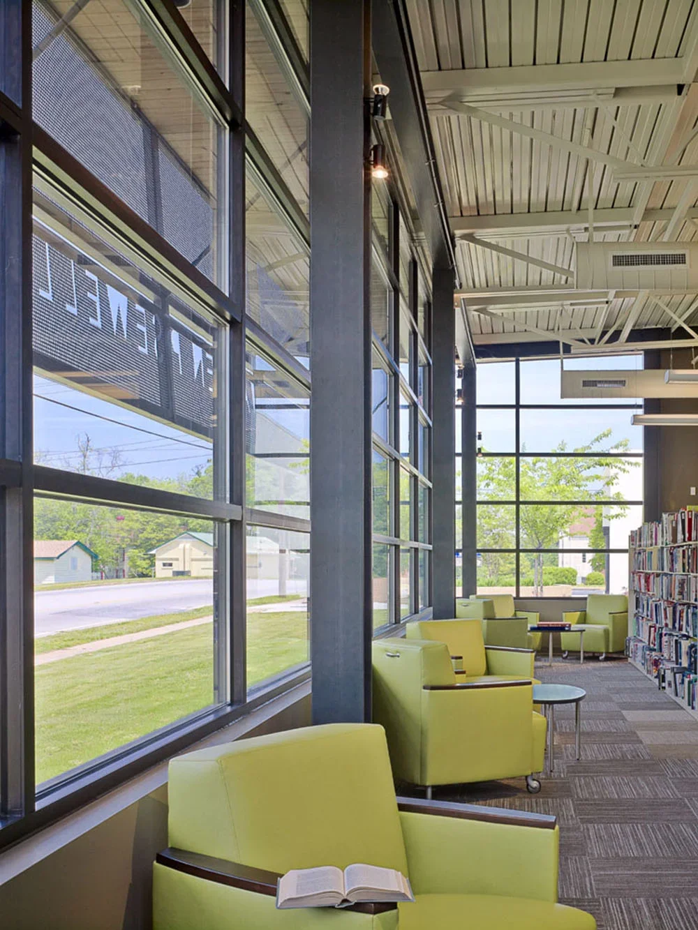 Interior of a modern library with large glass windows, lime green armchairs, a book on one chair, bookshelf on the right, and trees visible outside.
