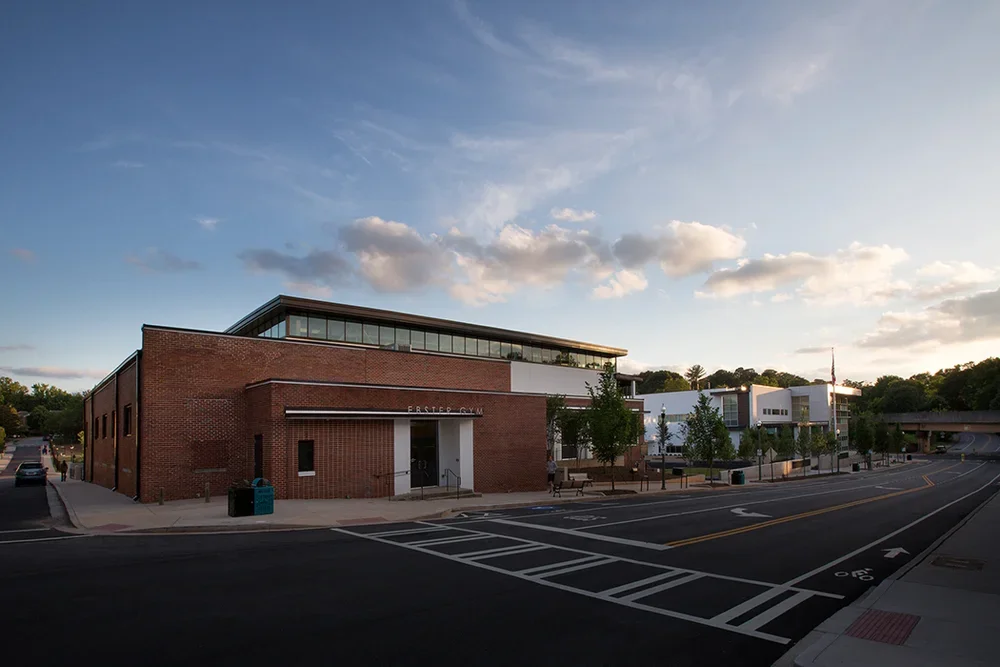 A modern building with a brick facade and large glass windows, located on a quiet street with trees and benches, during dusk with a partly cloudy sky.