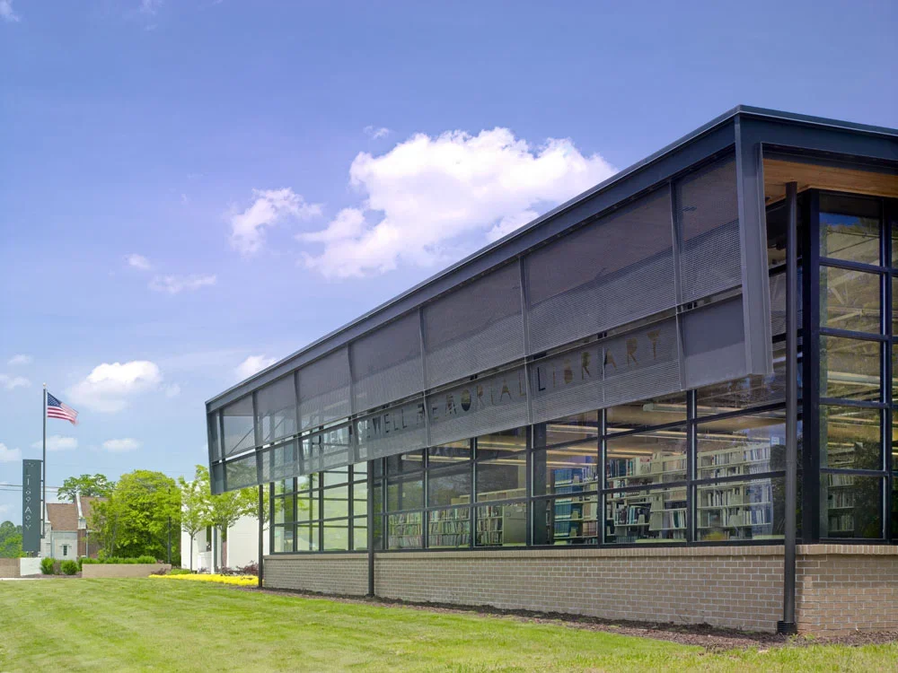 Modern library building with glass walls displaying shelves of books, surrounded by a well-kept lawn, blue sky, and an American flag in the background.