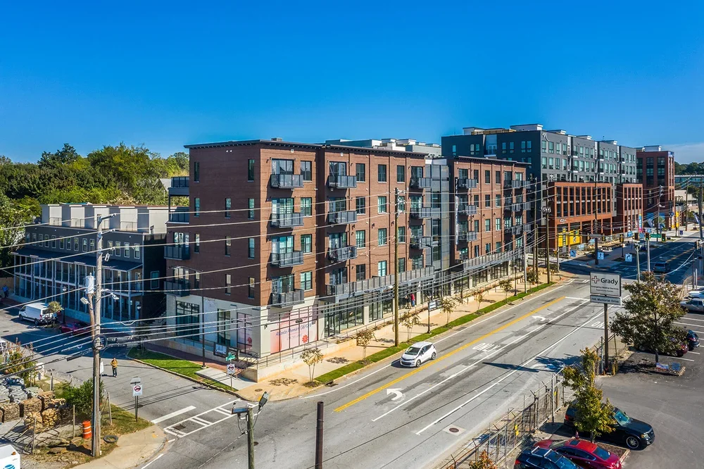 A modern multi-story apartment building under construction with several sections and balconies, situated along a busy city street with cars parked along the curb and mature trees lining the sidewalk.