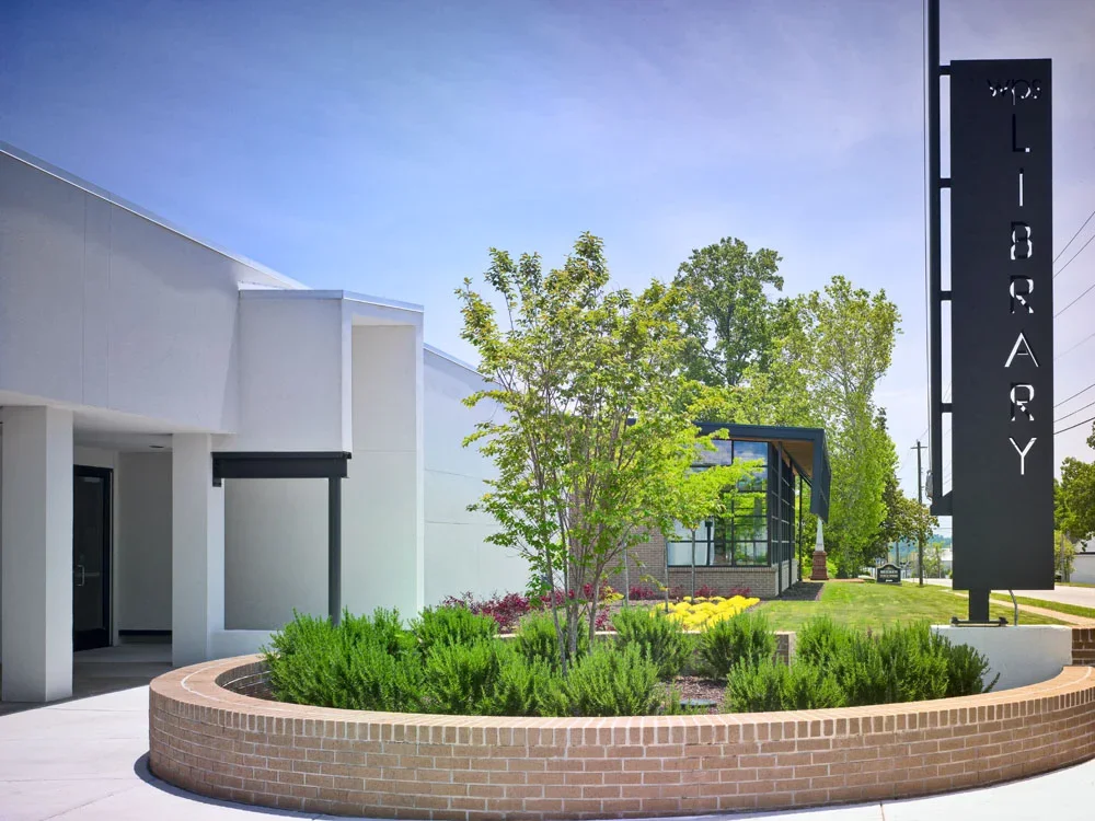 Exterior of a building with a large landscaped flower bed containing bushes and a small tree, and a tall sign that reads "Library" in vertical letters.