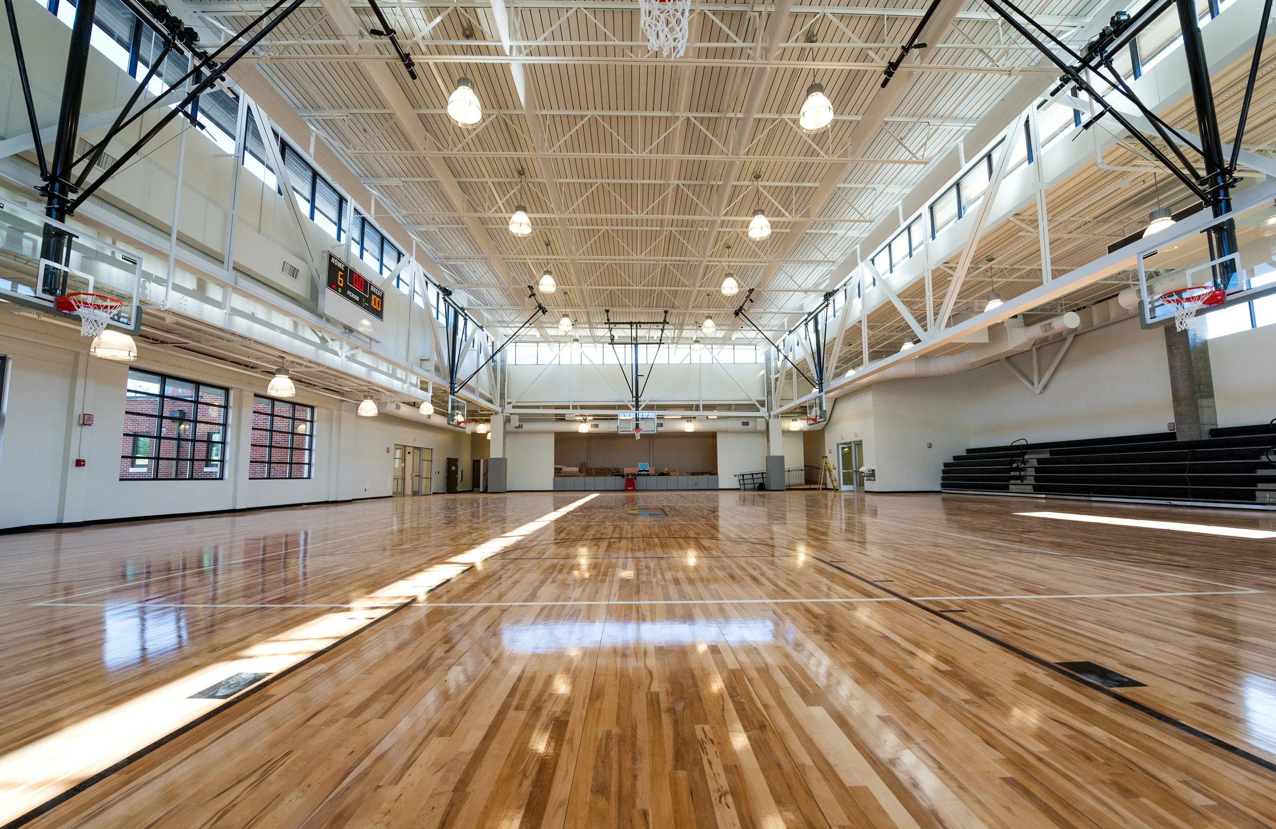 Empty indoor basketball court with polished wooden floor, basketball hoops, scoreboards, and high ceiling with bright lighting.