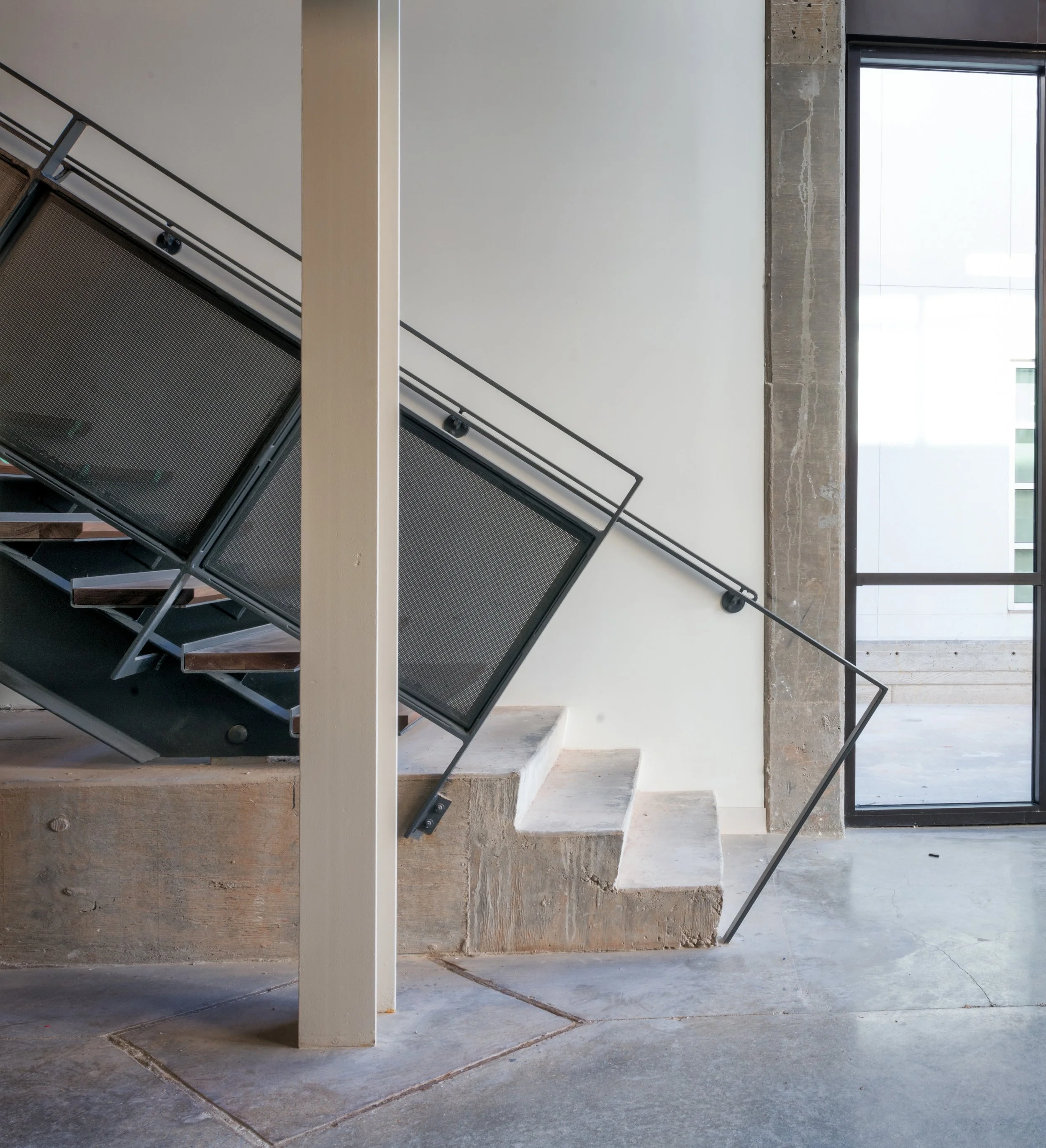 Interior of a modern building with concrete and white walls, stairs with black metal railings, and large windows letting in natural light.