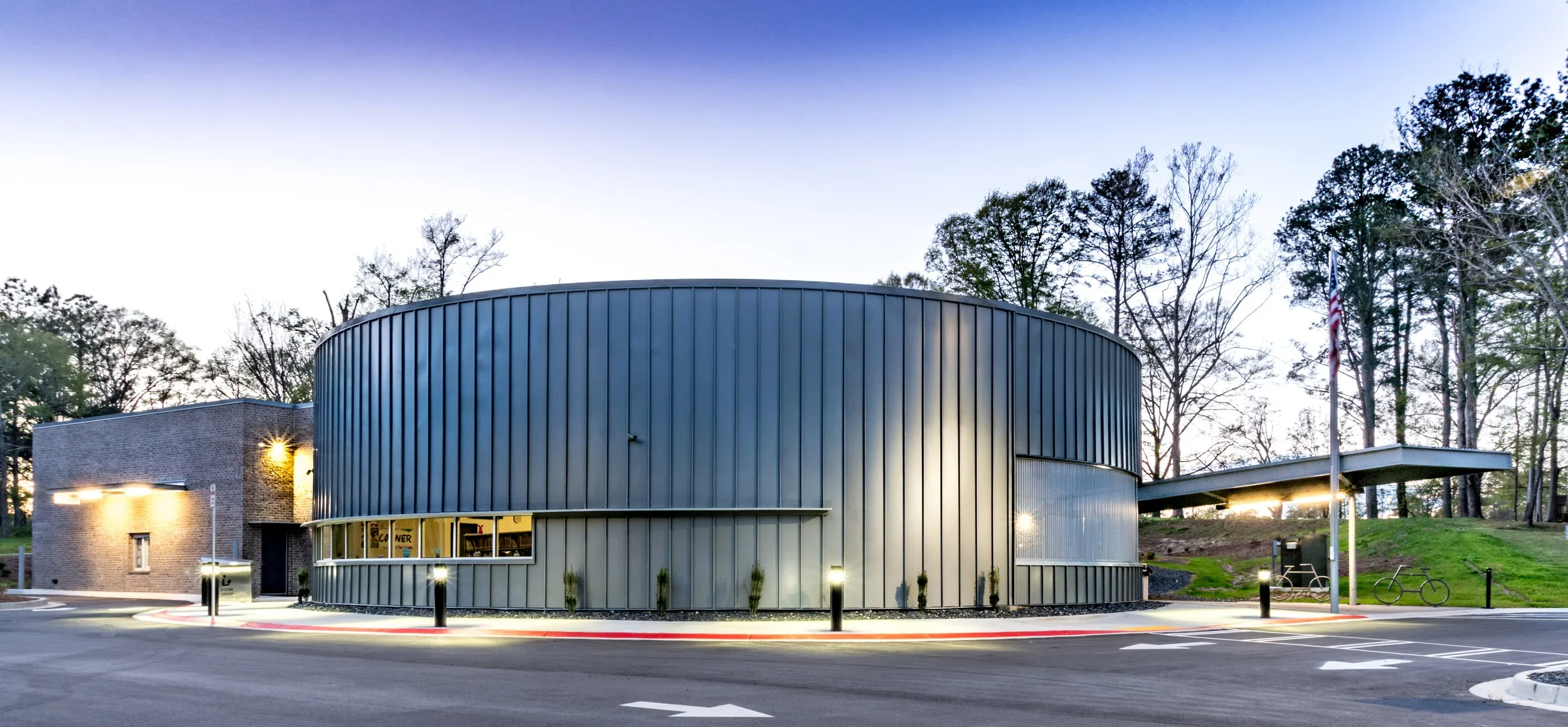 Modern building with curved metal siding and brick section, surrounded by trees and parking lot with American flag, bicycles, and streetlights at dusk.