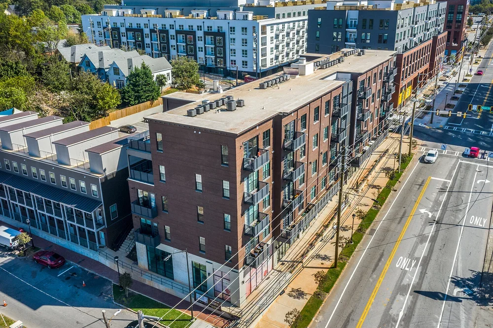 Aerial view of a modern apartment building with brick facade and balconies, situated along a busy street with traffic lights and cars, in an urban area with trees and smaller houses nearby.