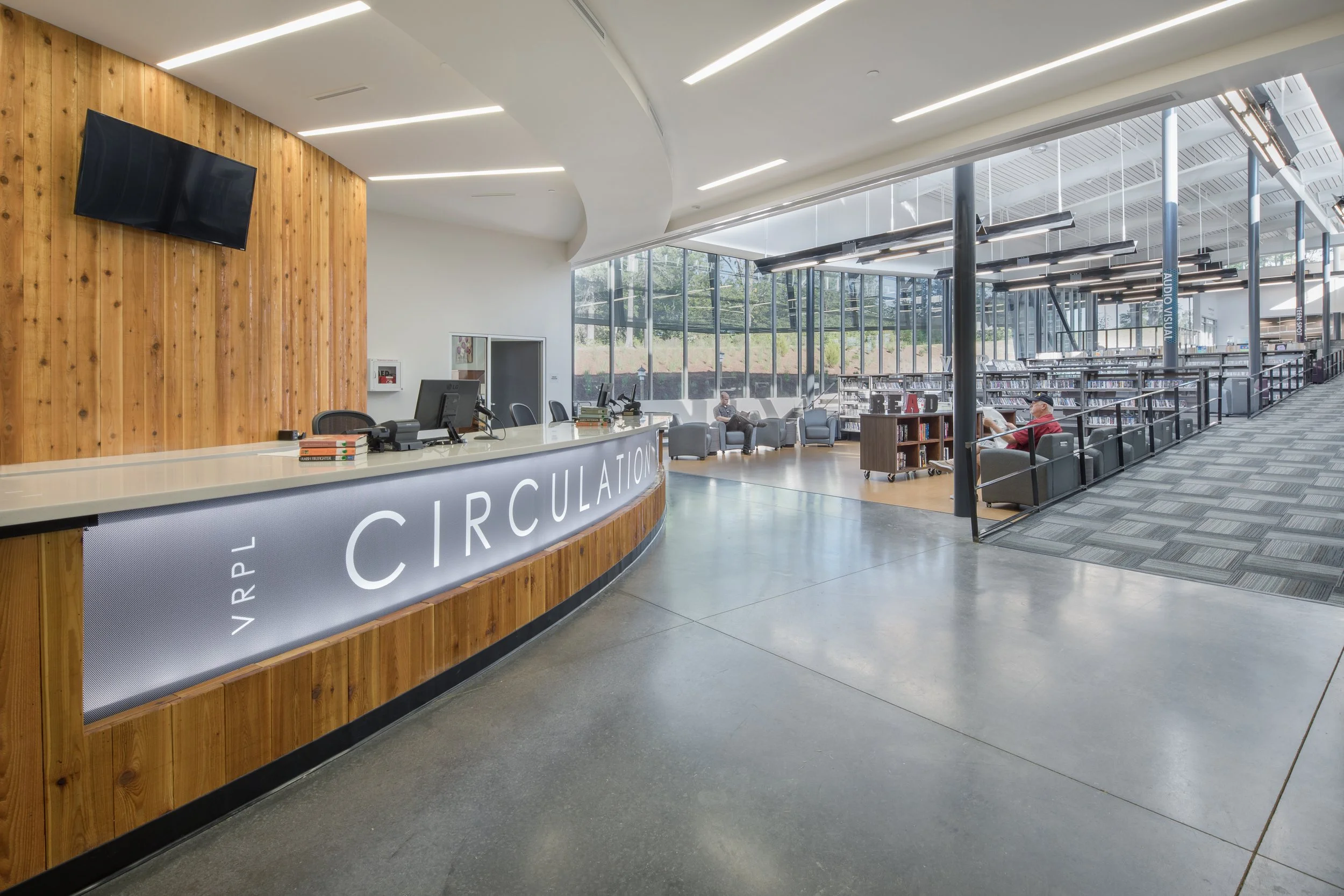 Modern library interior with a reception desk on the left and seating area with chairs and people reading books on the right, large floor-to-ceiling windows providing natural light, wooden accents, suspended ceiling lighting.