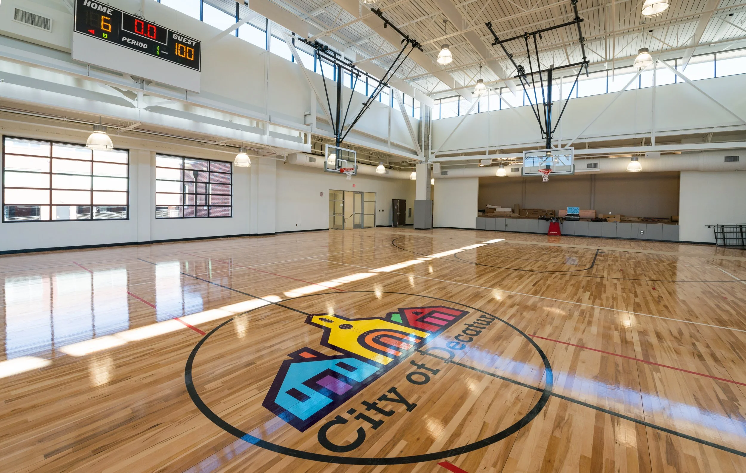 Interior of a modern indoor basketball gymnasium with a polished wood floor, basketball hoops, and a scoreboard showing 6 to 0 in the game, with 1 minute remaining in the first period. The gym has large windows and bright lighting.