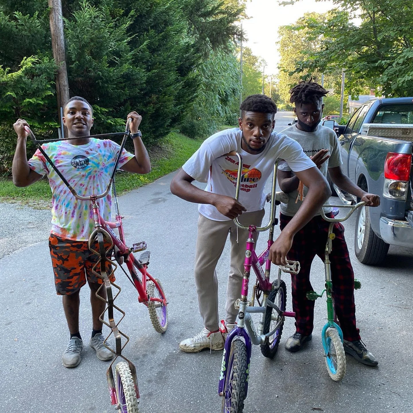 Three young men standing on a street with two of them holding small colorful bicycles, one with a pink bike and another with a purple bike, while the third stands behind them wearingCamouflage shorts and a tie-dye shirt.
