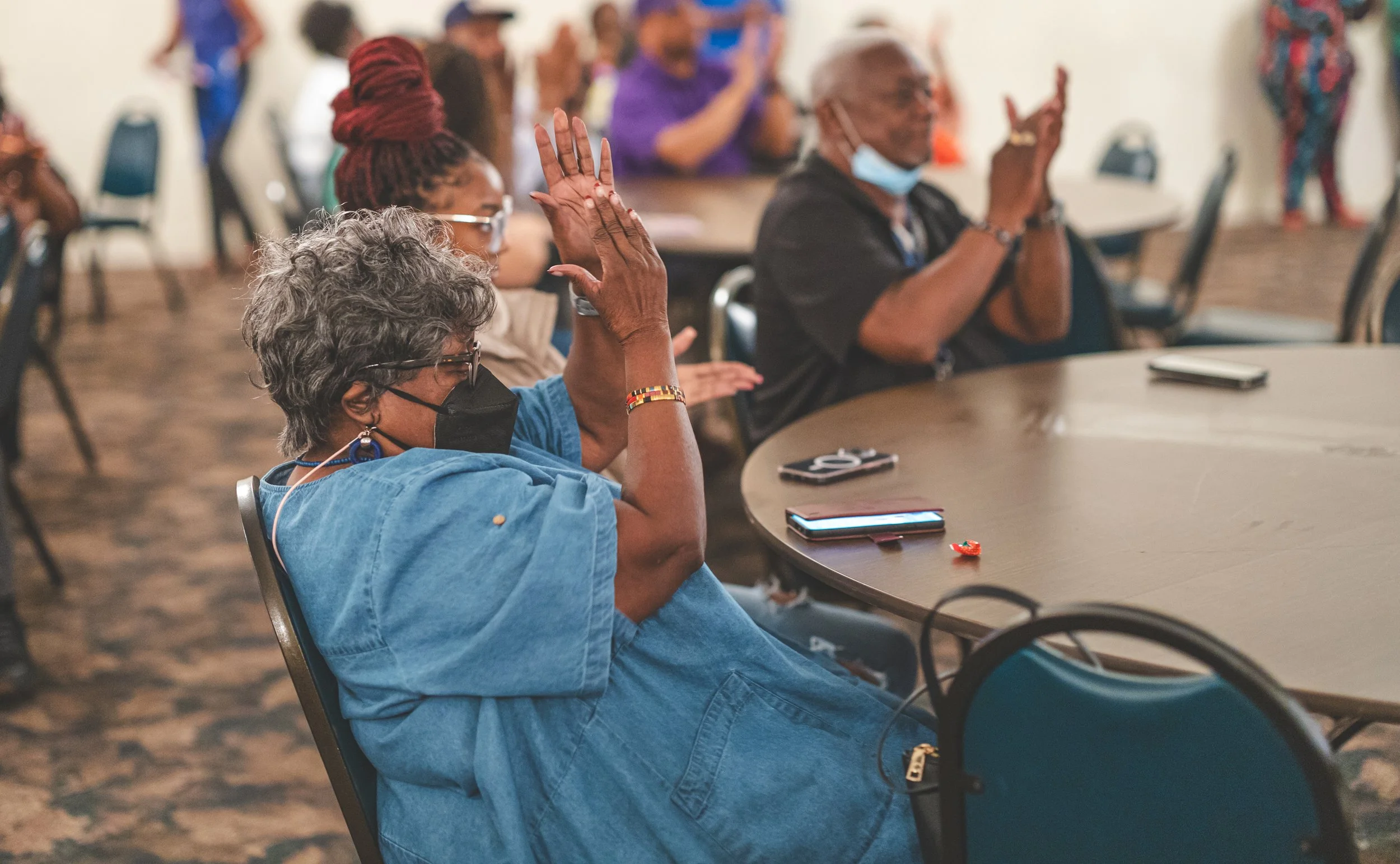 People sitting at a round table during a meeting or conference, raising their hands to participate, with some wearing masks.