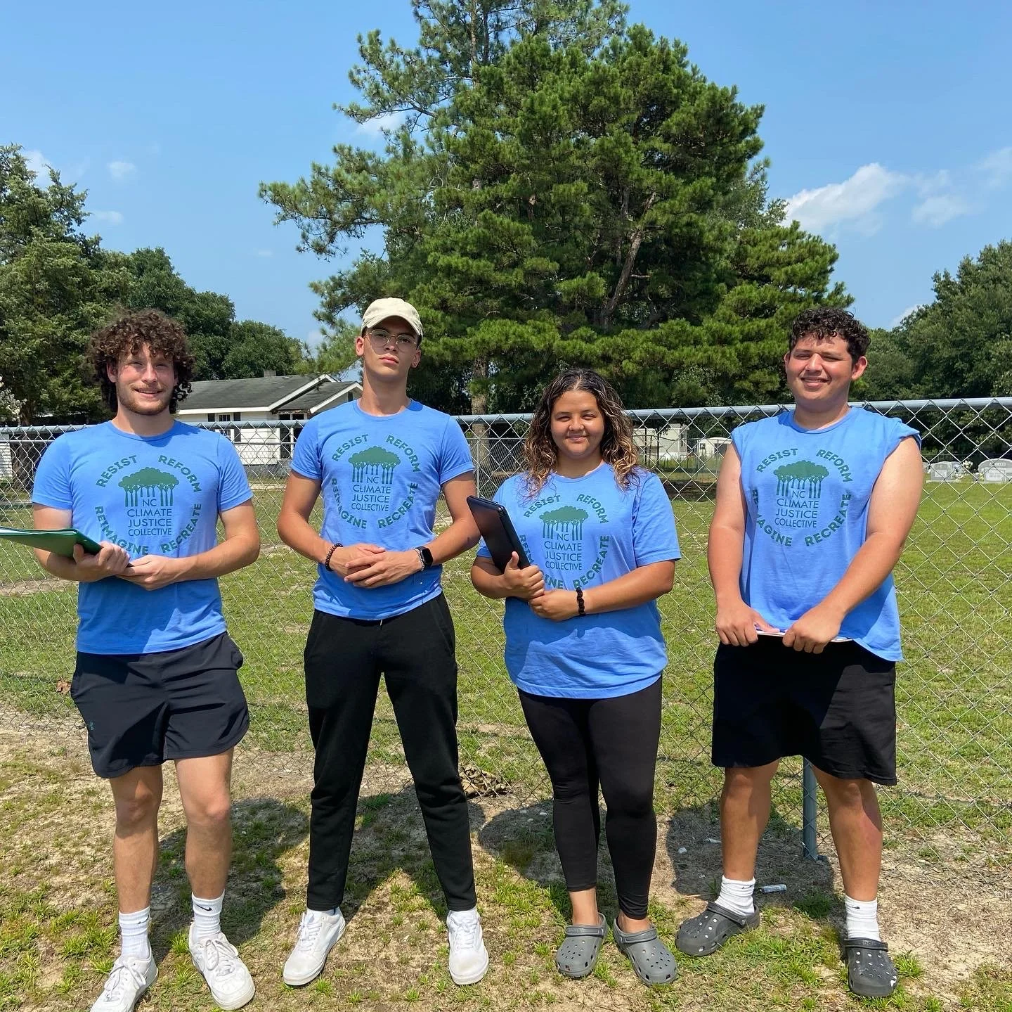 Four young people standing outdoors in front of a chain-link fence, wearing matching blue T-shirts with a climate justice message, on a sunny day with trees and a house in the background.