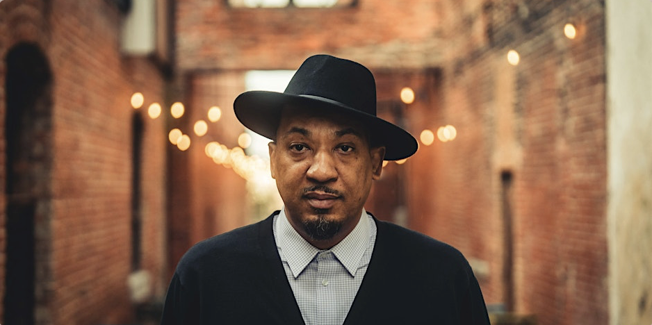 A man wearing a black hat and a dark sweater over a light checked shirt, standing in an alley with brick walls and string lights.