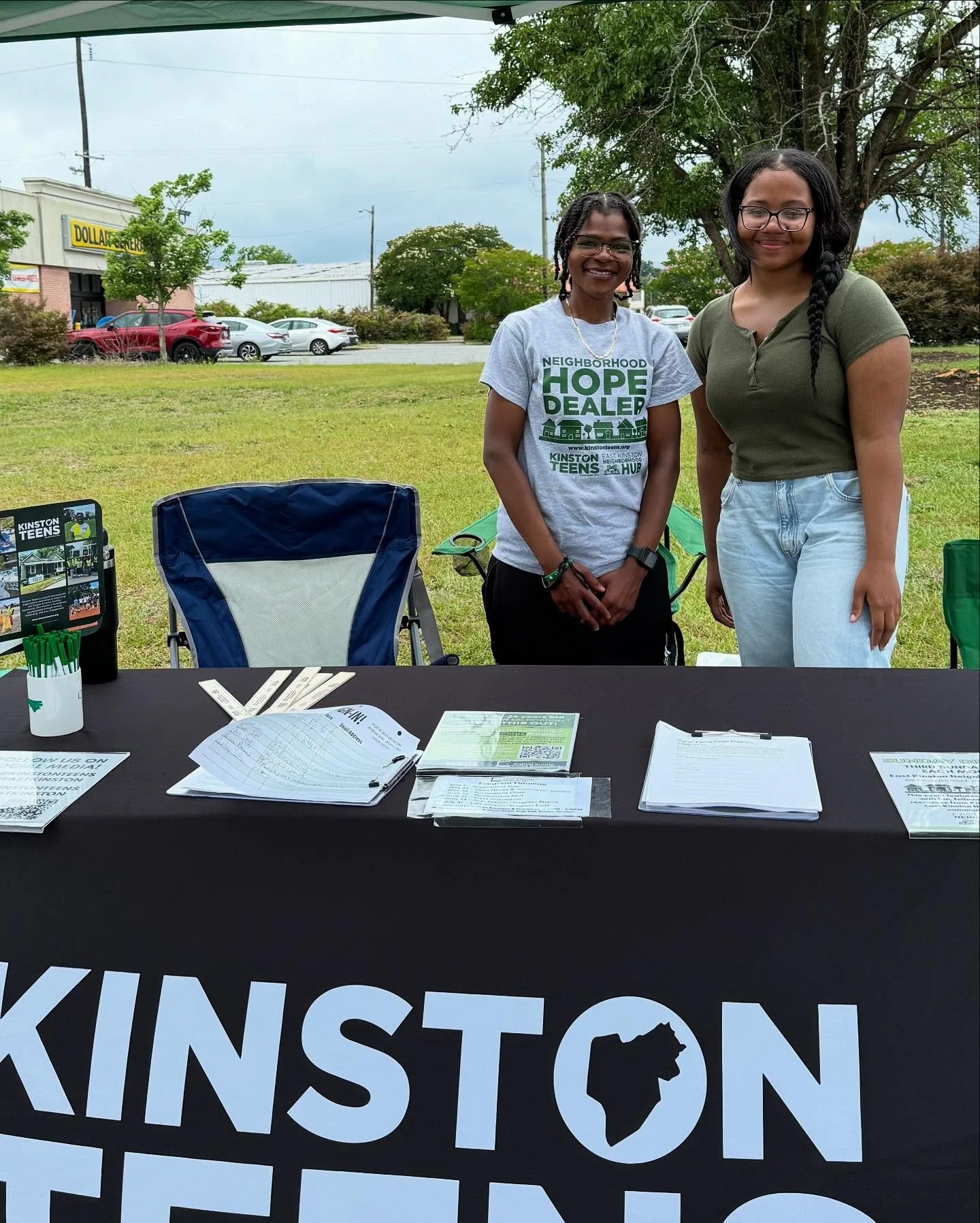 Two women standing behind a table at an outdoor event with informational flyers and pens, in a grassy area with trees and cars in the background.