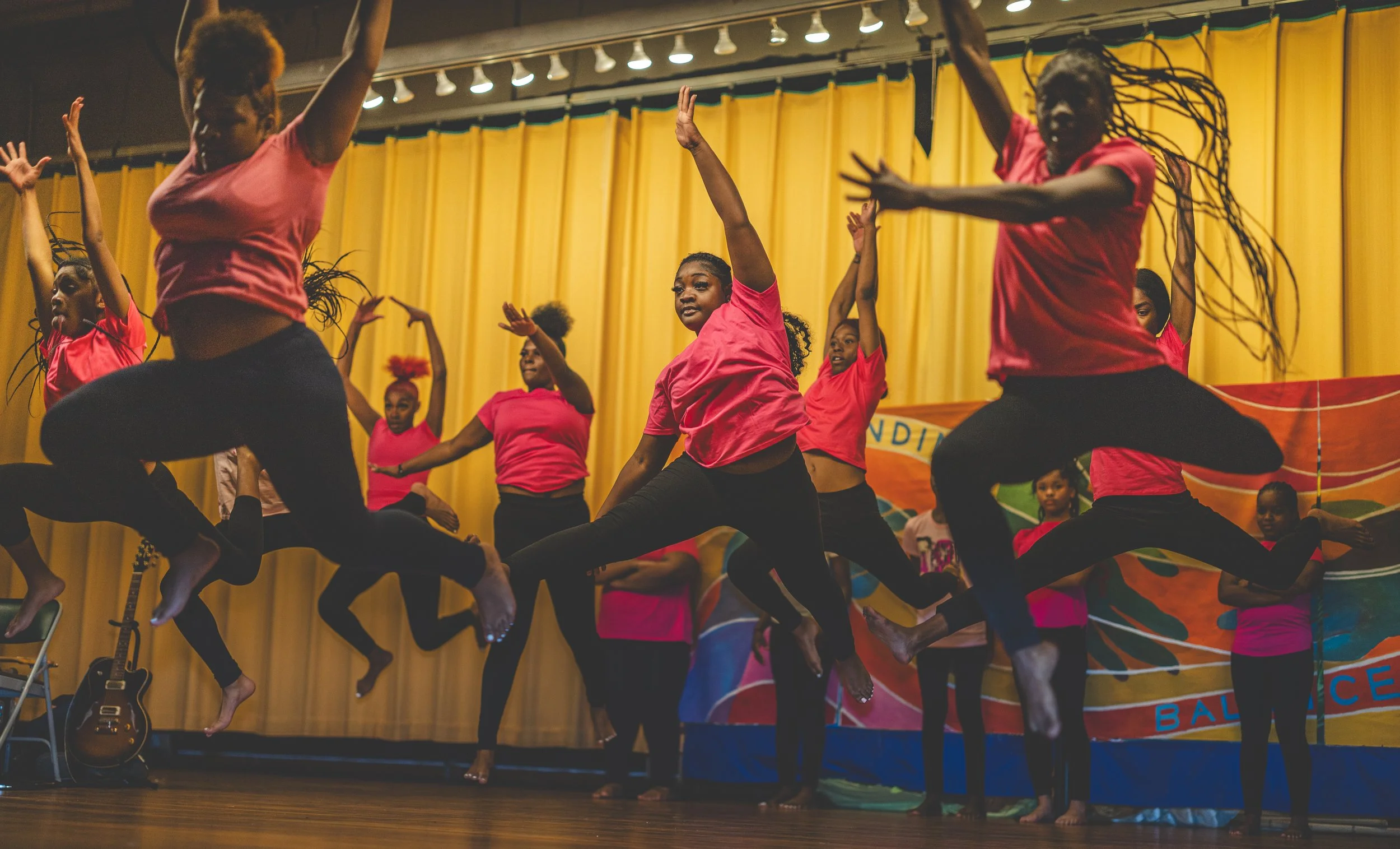 Group of young women performing a dance on stage with yellow curtains in the background, wearing pink shirts and black pants, some jumping with arms raised.