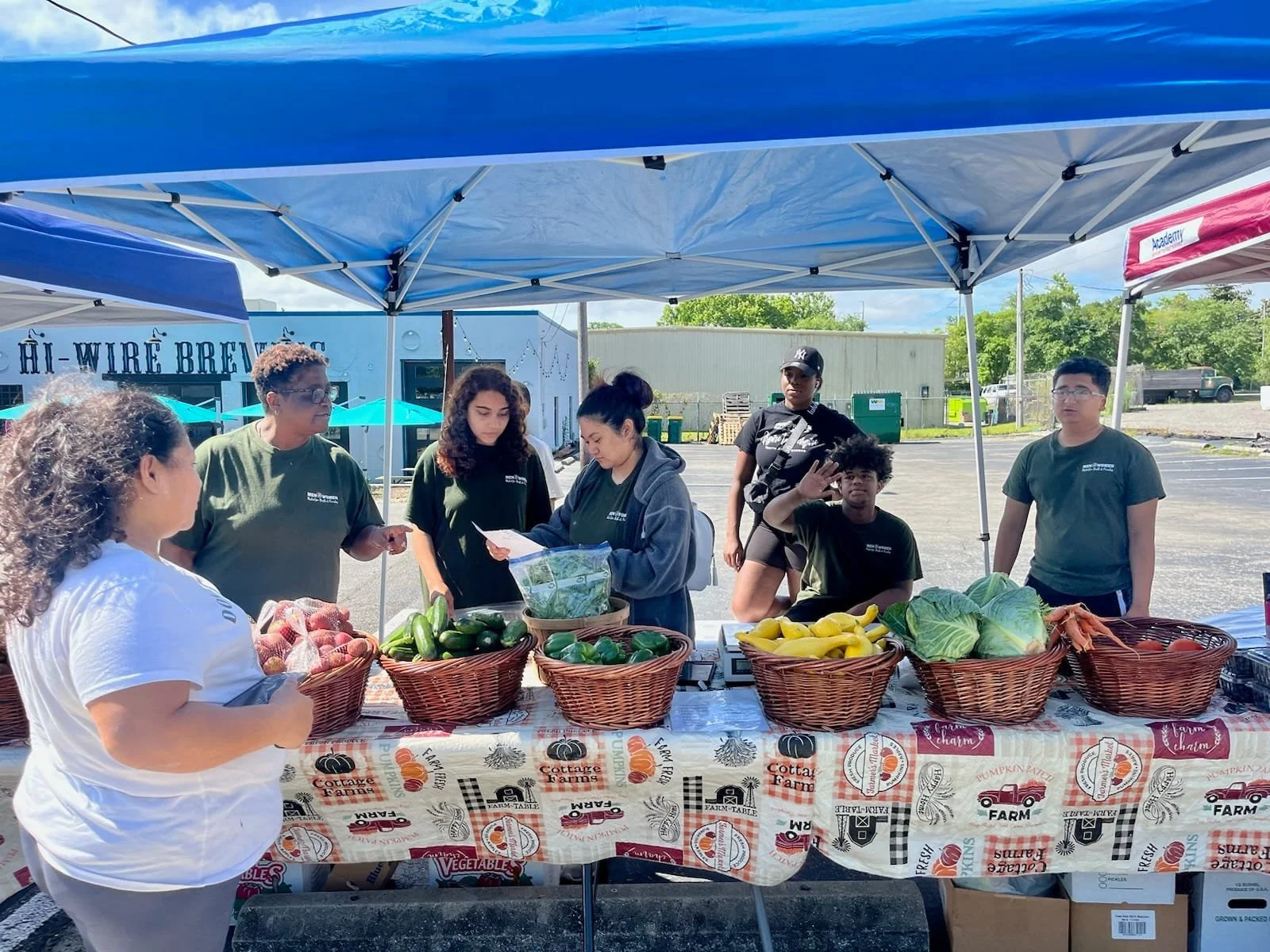 A group of people at an outdoor farmers market stand under a blue canopy, with baskets of fresh vegetables like zucchini, cabbage, and yellow squash on a table covered with a farm-themed tablecloth, and a white building in the background.