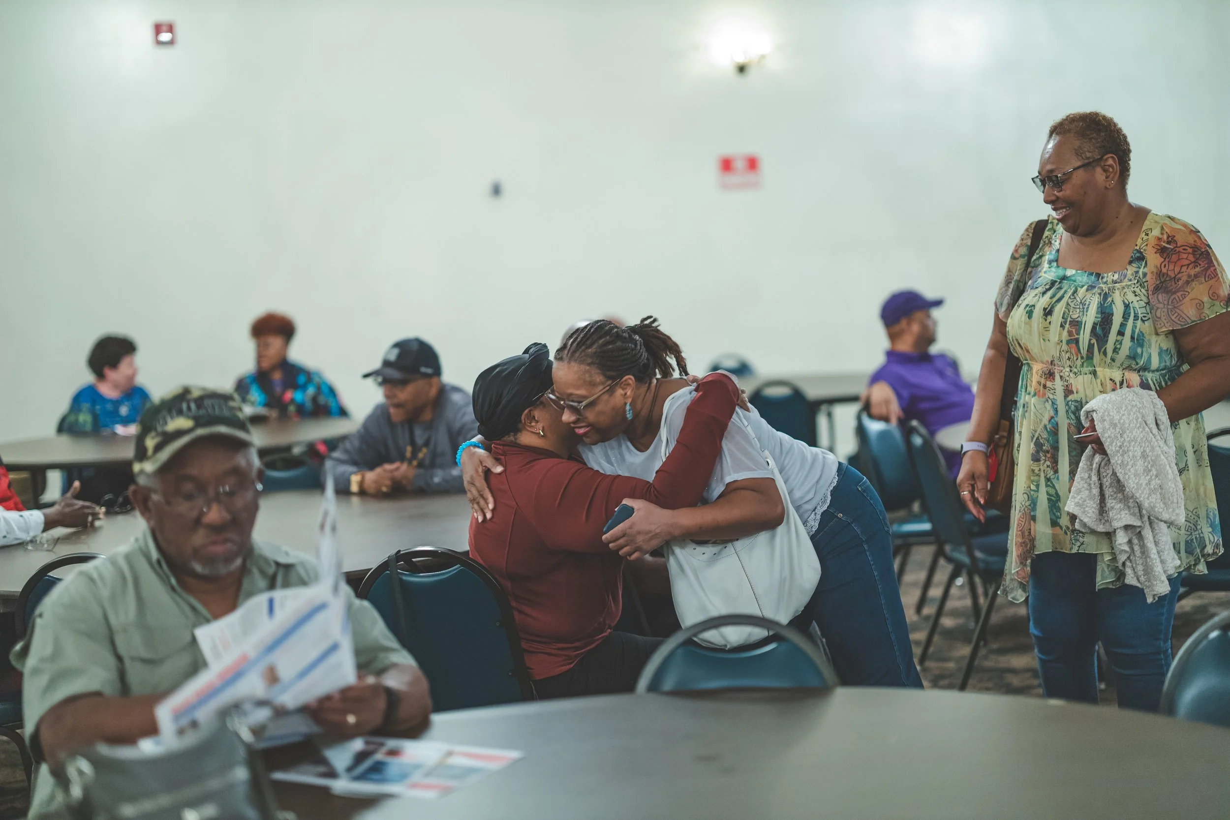 People hugging and smiling in a gathering, with some seated at tables and others standing, in a large room.