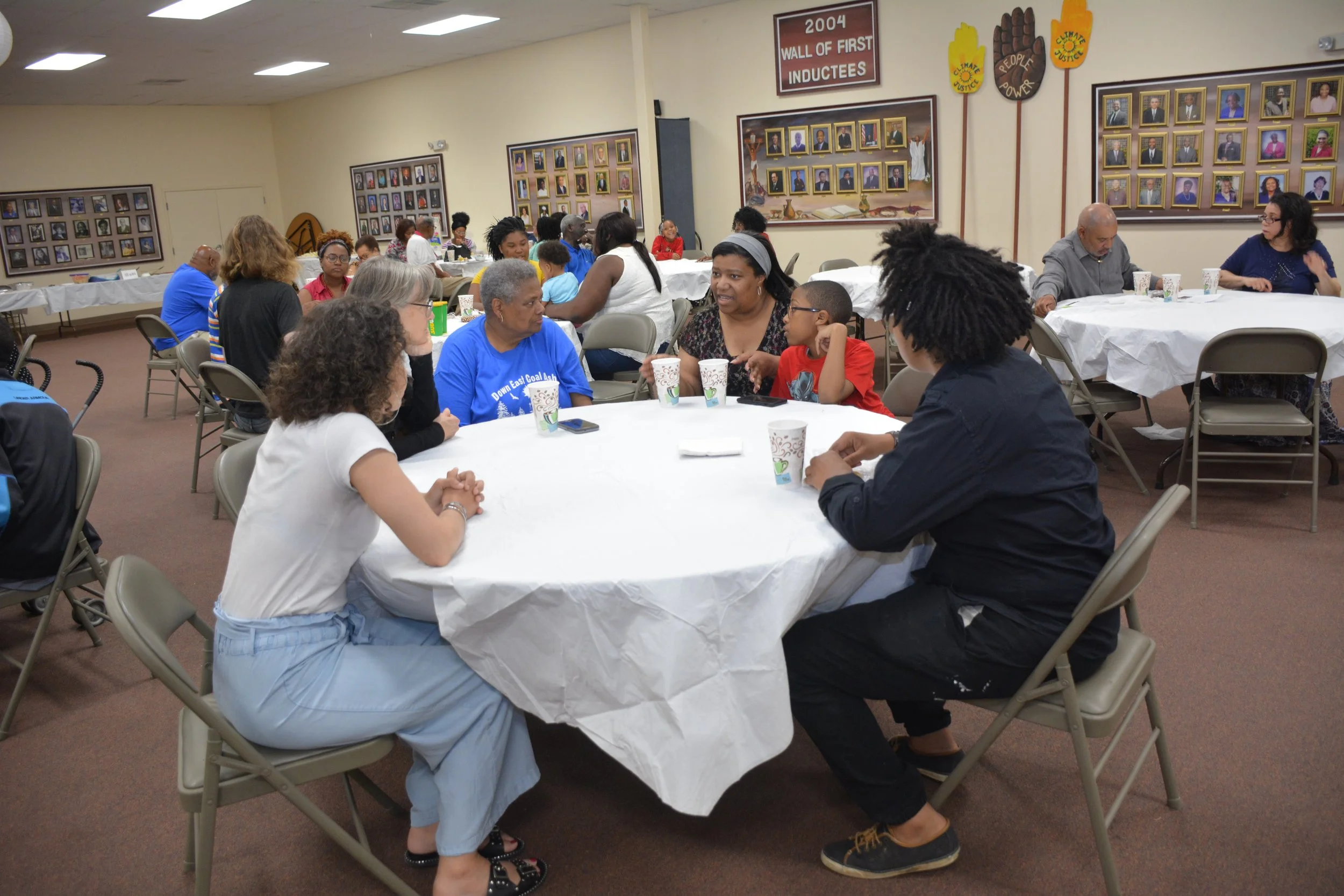 People sitting at round tables engaged in conversation, in a community gathering hall with photo collages and plaques on the walls.