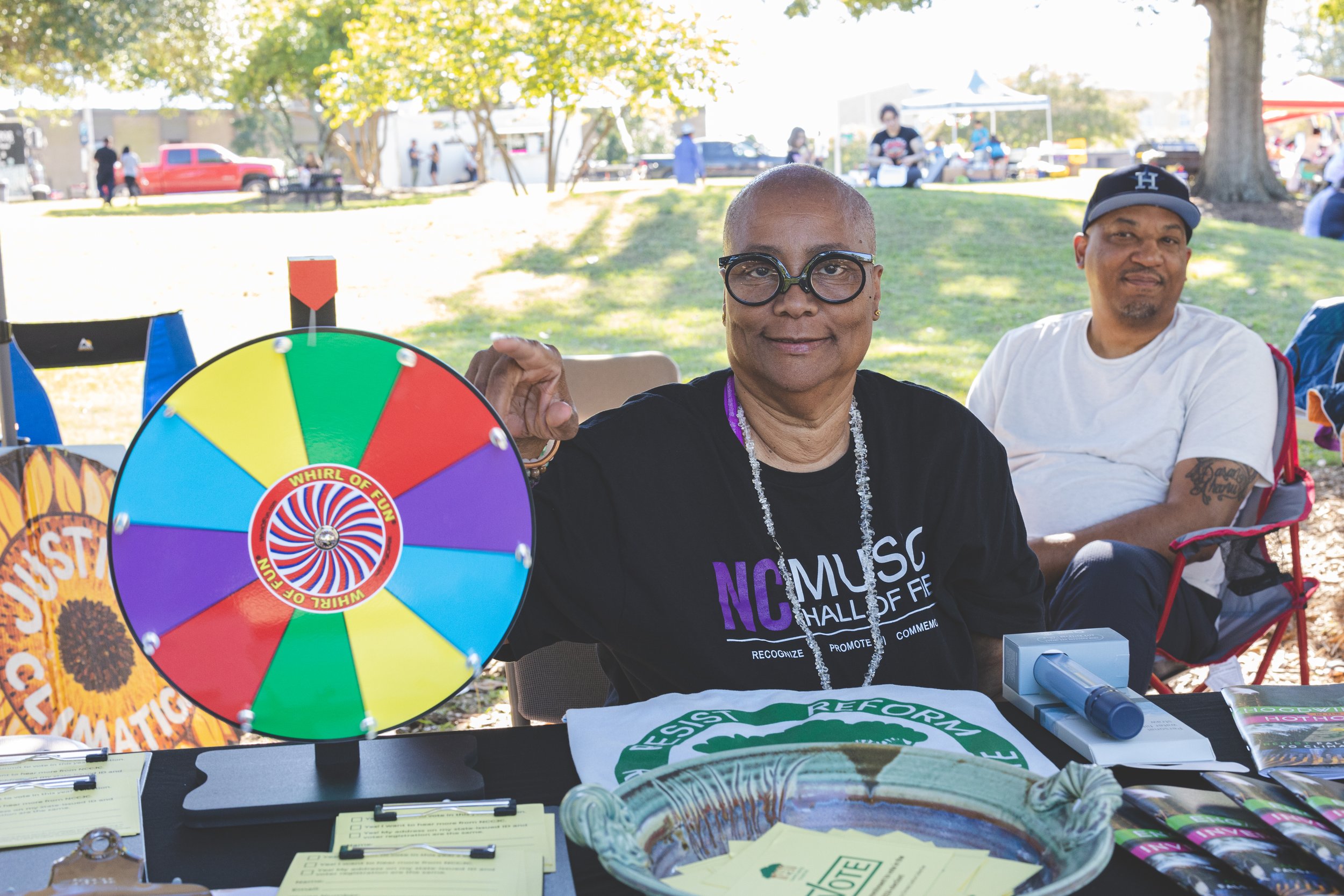 A woman with short hair and glasses holding a colorful spinning wheel labeled 'Whirl of Fun' at an outdoor event with tents and trees in the background. She sits at a table with pamphlets and a bowl, and a man with a cap is sitting behind her.