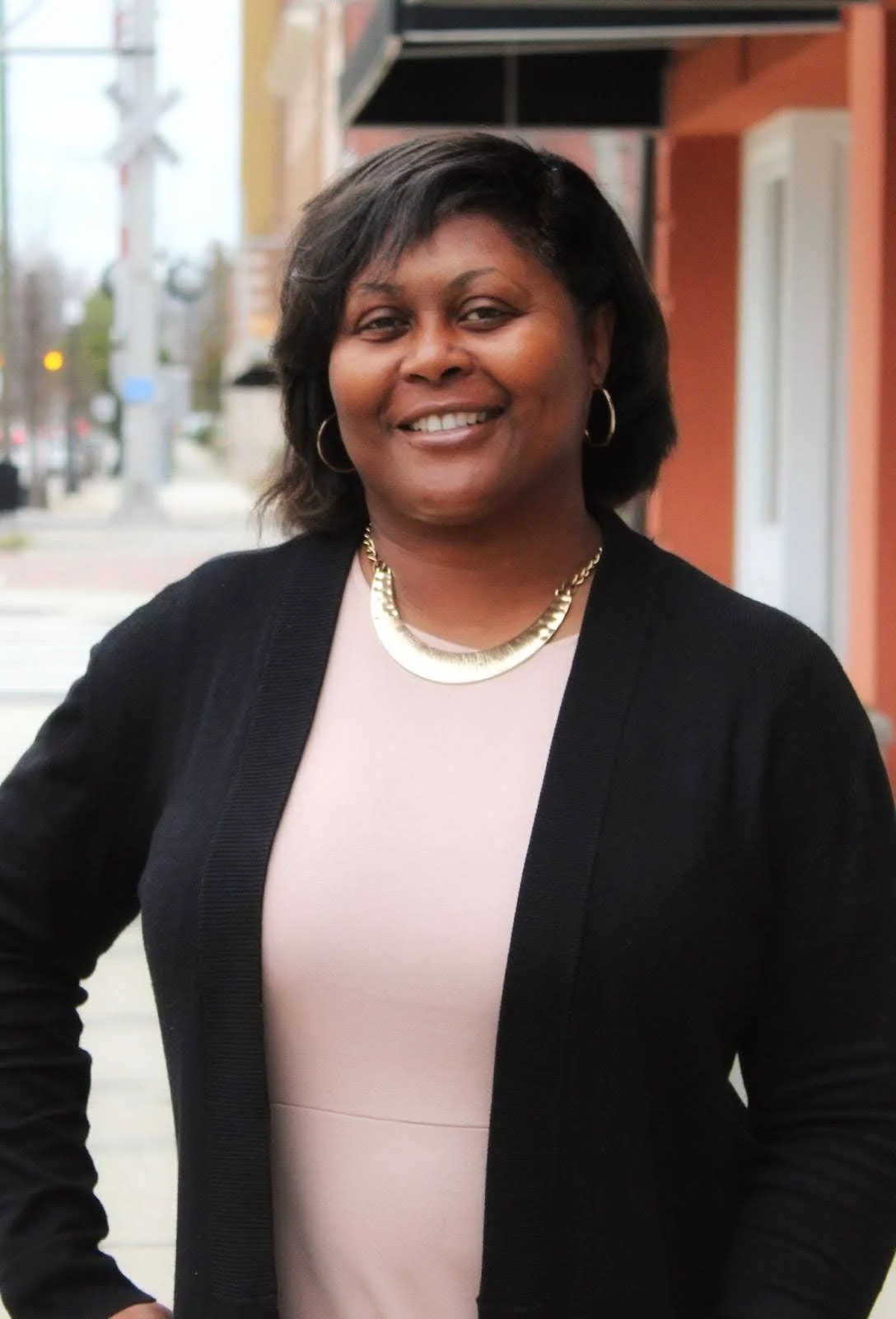 Portrait of an African American woman smiling outdoors on a city sidewalk, wearing a light pink top, black cardigan, gold necklace, and hoop earrings.