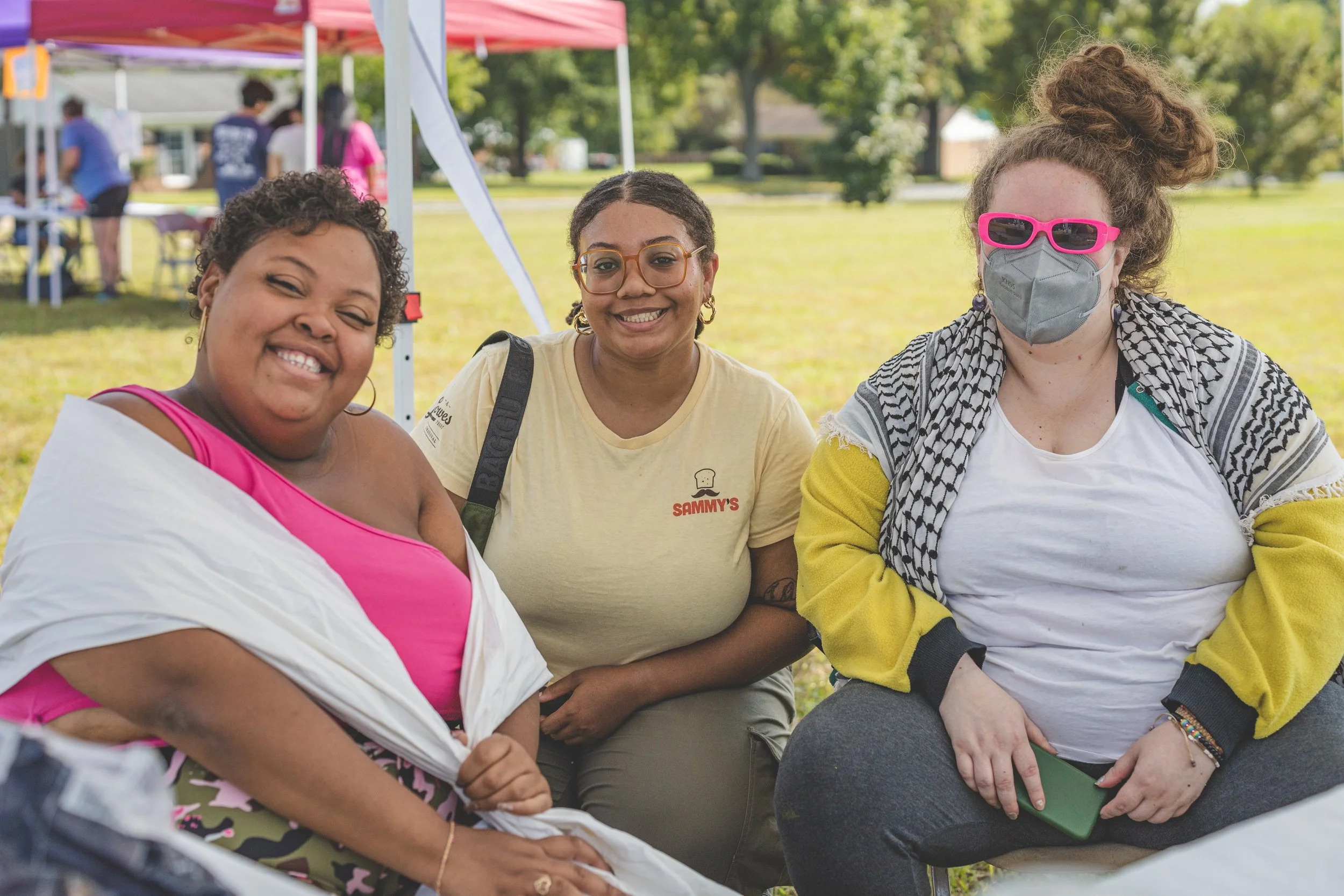 Three women sitting outdoors at a park event, smiling at the camera. The woman on the left has short curly hair and is wearing a pink top with a white shawl. The woman in the middle has glasses, earrings, and is wearing a yellow t-shirt with a logo t