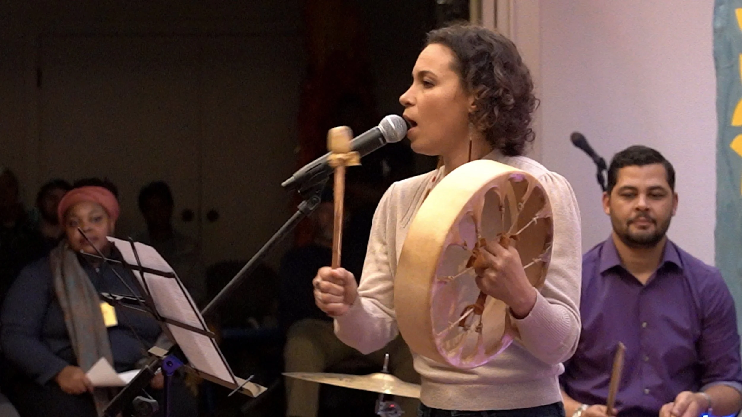 A woman singing into a microphone while playing a frame drum, accompanied by a man playing percussion on the right and another person with sheet music and mallet in the background.