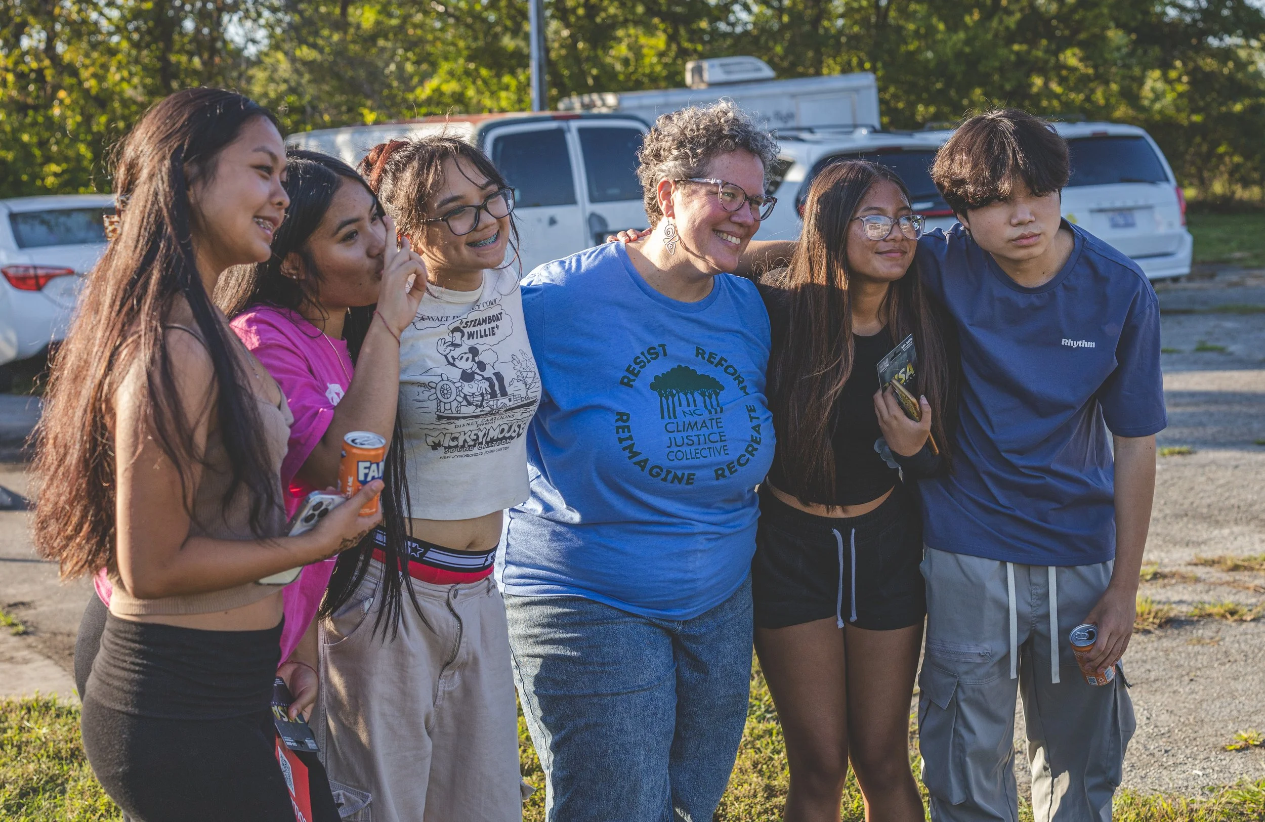 Group of six diverse young people and an older woman standing outdoors, smiling with their arms around each other, during daytime.