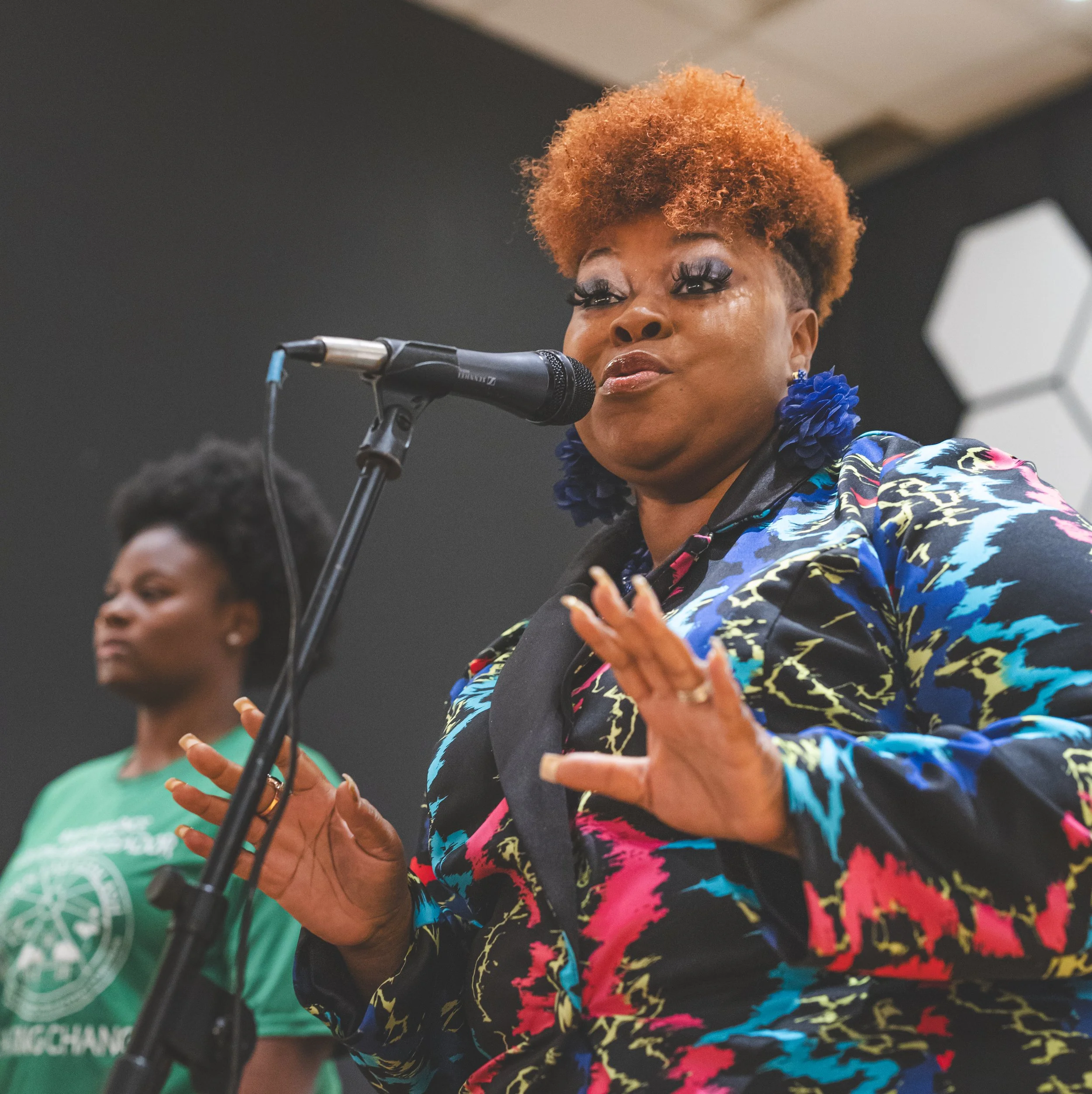 A woman with short, curly, reddish-brown hair speaking into a microphone in an indoor setting. She is wearing large blue floral earrings and a colorful, patterned jacket. Another woman with short, natural black hair wearing a green shirt stands in th