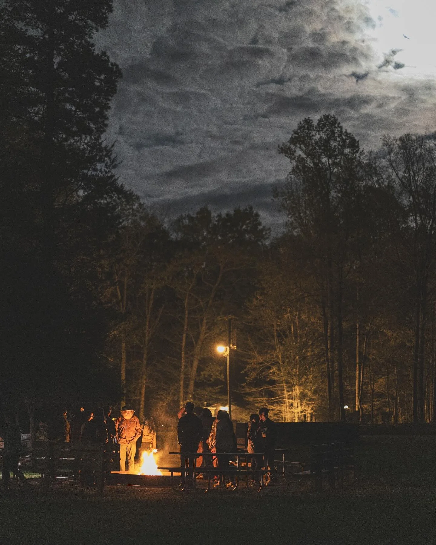 People gathered around a campfire at night in a wooded area with trees and dark clouds in the sky.
