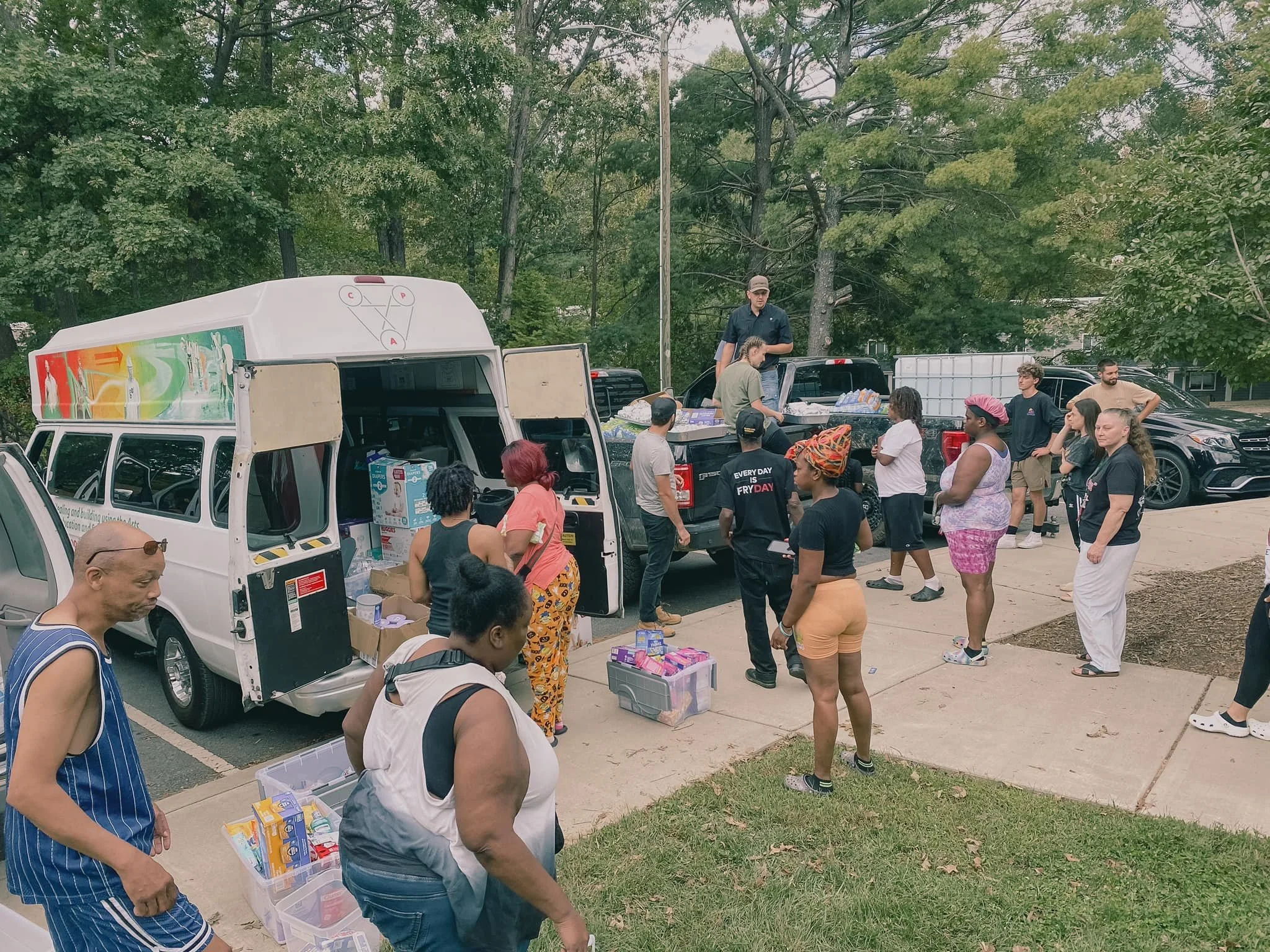 People gathered around trucks and a van, receiving and distributing supplies, outdoors in a park or community area with trees in the background.