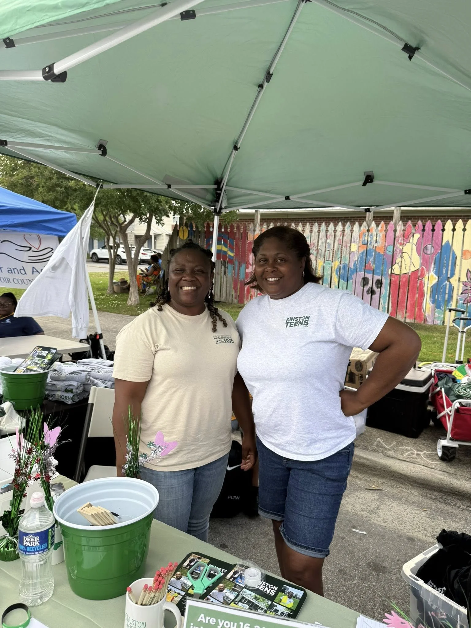 Two women standing underneath a green canopy at an outdoor event, smiling for the camera. There are tables with items and a colorful painted fence in the background.