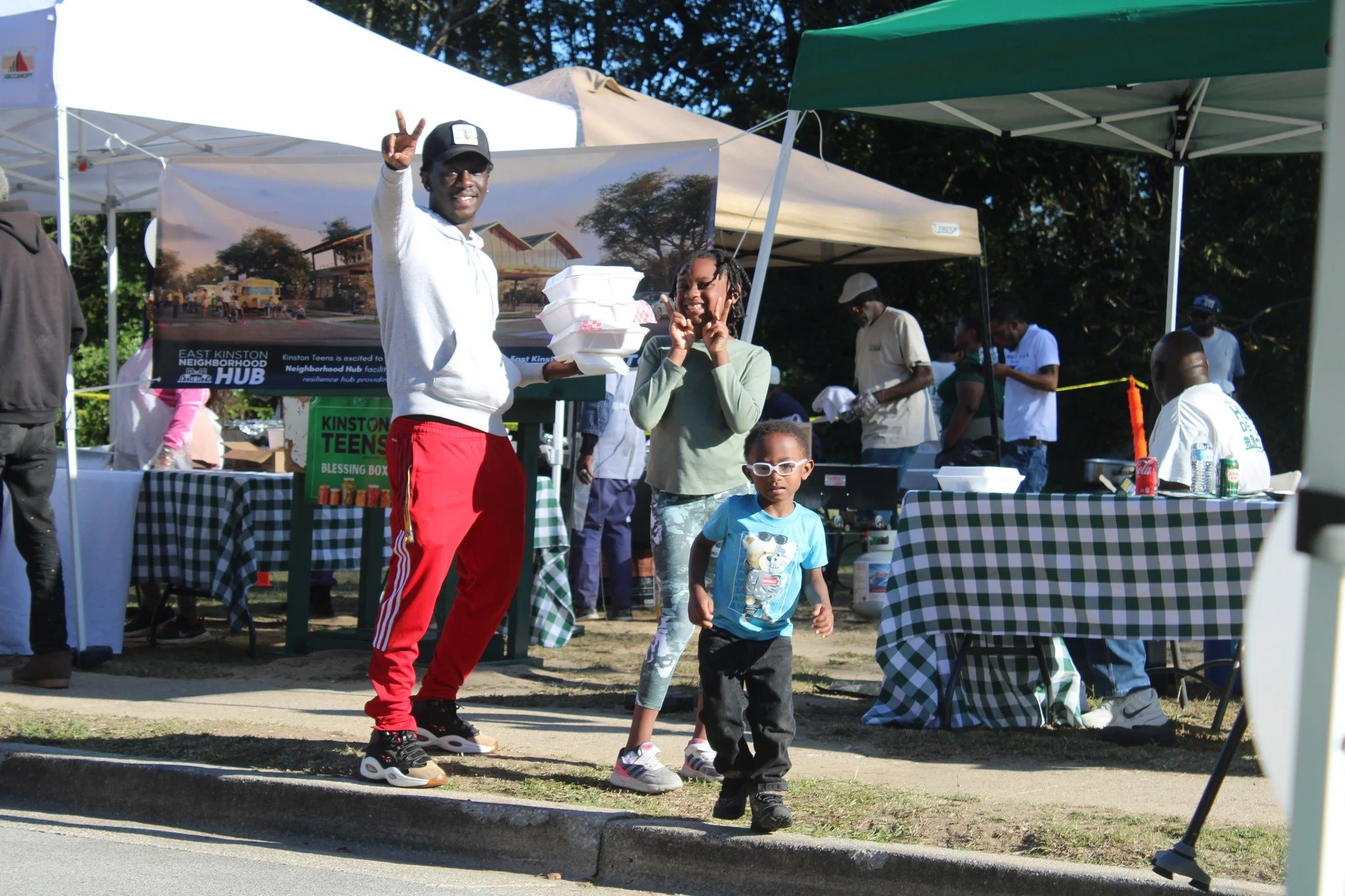 People at an outdoor community event, with a man giving a peace sign, a woman and a young boy in front, and tents in the background.