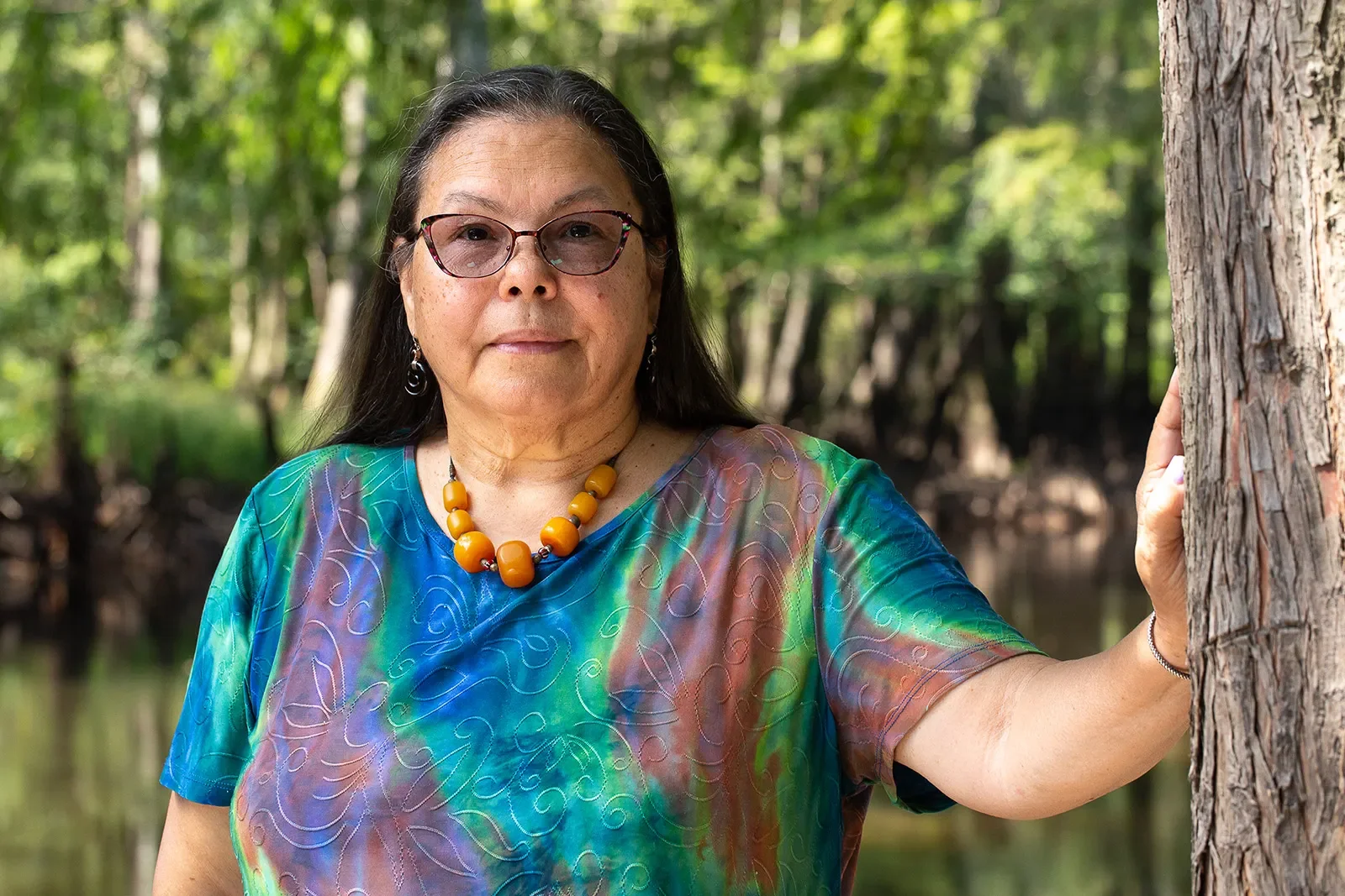 Portrait of a woman with glasses and long dark hair, wearing a colorful blouse and yellow beaded necklace, standing by a tree in a forested area with a lake in the background.