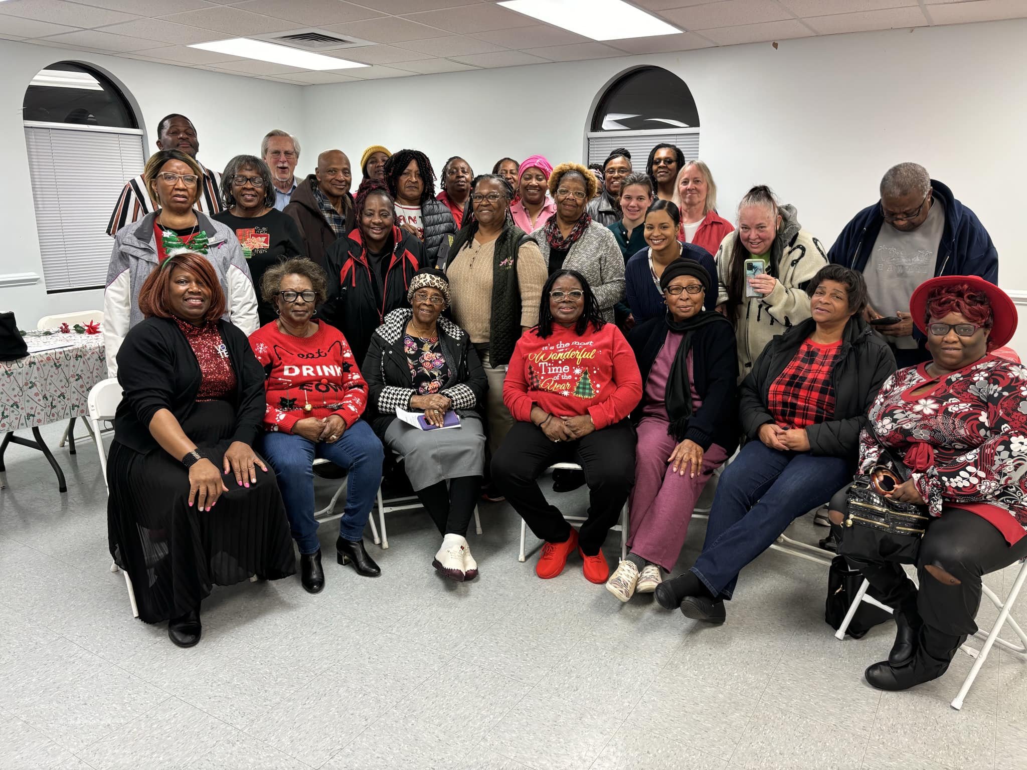 Group of diverse women and men gathered indoors for a holiday celebration, many wearing Christmas-themed clothing.